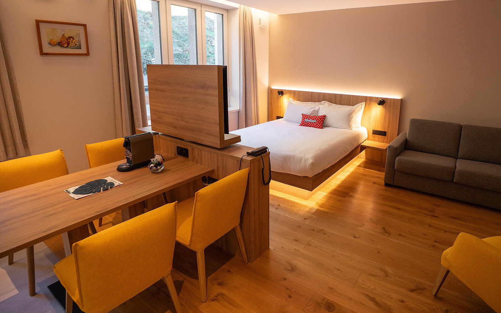 Ein modernes Hotelzimmer mit einem großen Bett, einem grauen Sofa und einem Holztisch, der von gelben Stühlen umgeben ist. Eine halbhohe Holzwand mit integriertem Fernseher trennt den Schlafbereich vom Essbereich. Warme Beleuchtung und Holzboden schaffen eine gemütliche Atmosphäre.