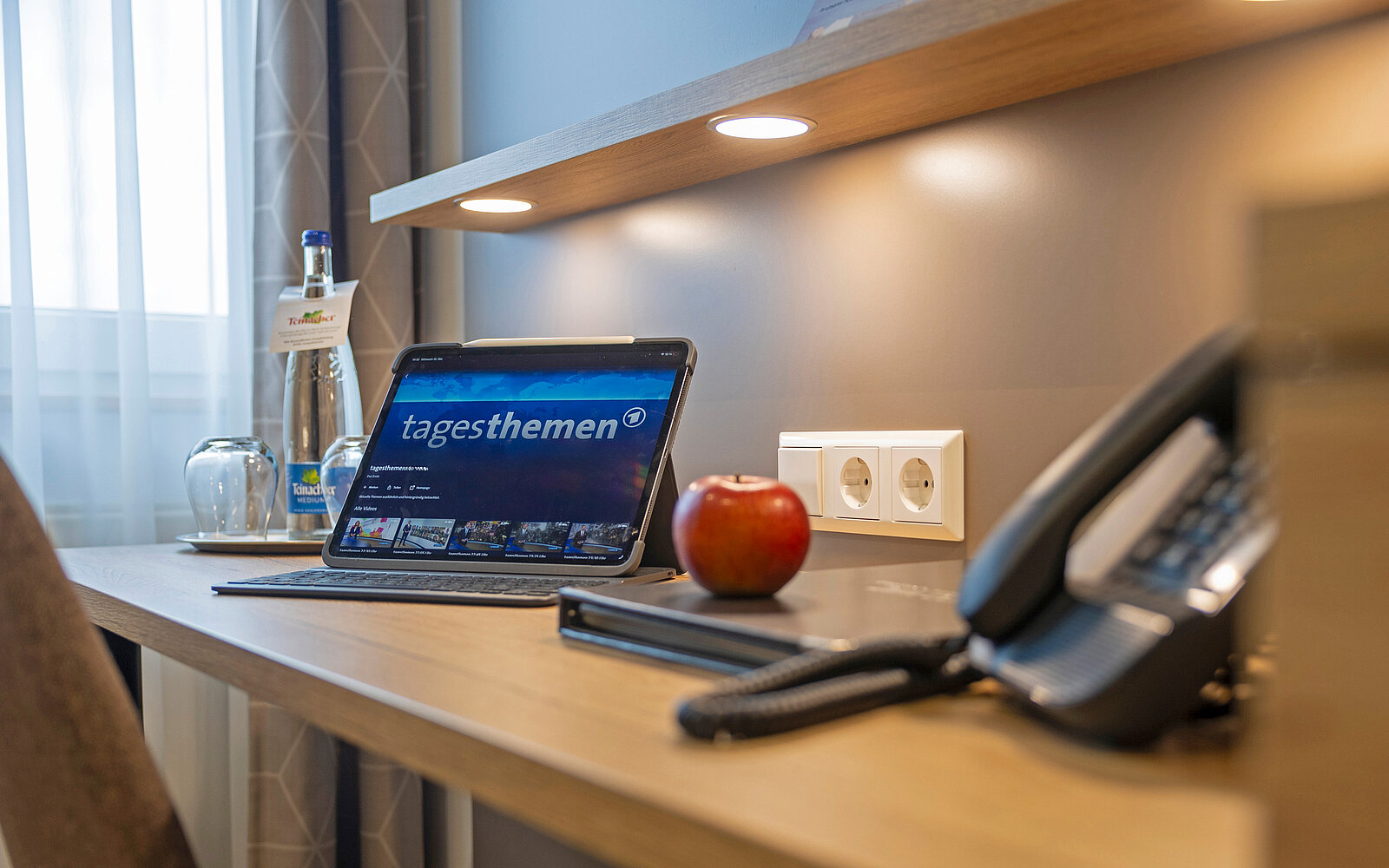 Ein Schreibtisch in einem Hotelzimmer zeigt ein Tablet mit der Webseite der "Tagesthemen". Daneben stehen eine Wasserflasche, ein Glas, ein roter Apfel und ein Telefon.