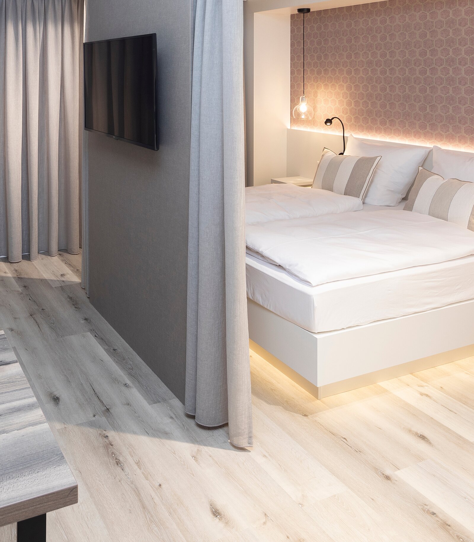 Ein modernes Schlafzimmer mit einem großen Bett, das von einer grauen Wand abgetrennt ist. An der Wand hängt ein Fernseher. Eine warme Beleuchtung und elegante Vorhänge tragen zur gemütlichen Atmosphäre bei. Der Boden ist aus hellem Holz.