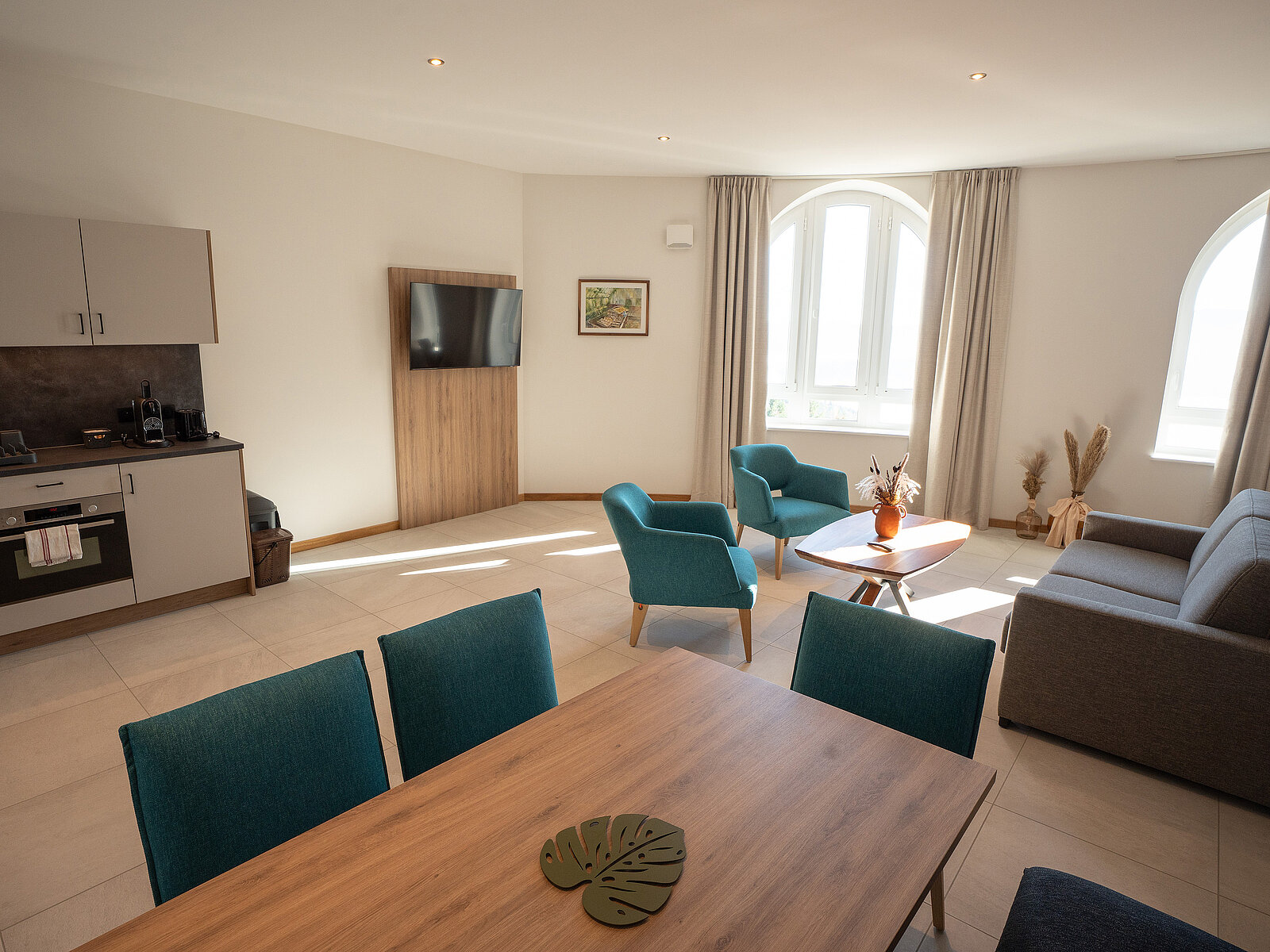 Ein modern eingerichtetes Wohnzimmer mit einem Esstisch im Vordergrund, zwei blauen Sesseln, einem Sofa, einem Flachbildfernseher an der Wand und einer kleinen Küchenzeile links. Große Fenster lassen viel Tageslicht herein, dekoriert mit Vorhängen.