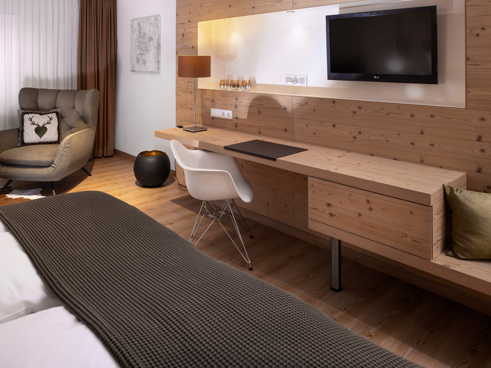 zieflekoch musterzimmer zulux, modernes Zimmer mit Holzdekor, wandmontiertem Fernseher, Schreibtisch mit Stuhl und anschliessender Sitzbank, gemütlichem Sessel und einem Bett mit dunkler Strukturdecke.