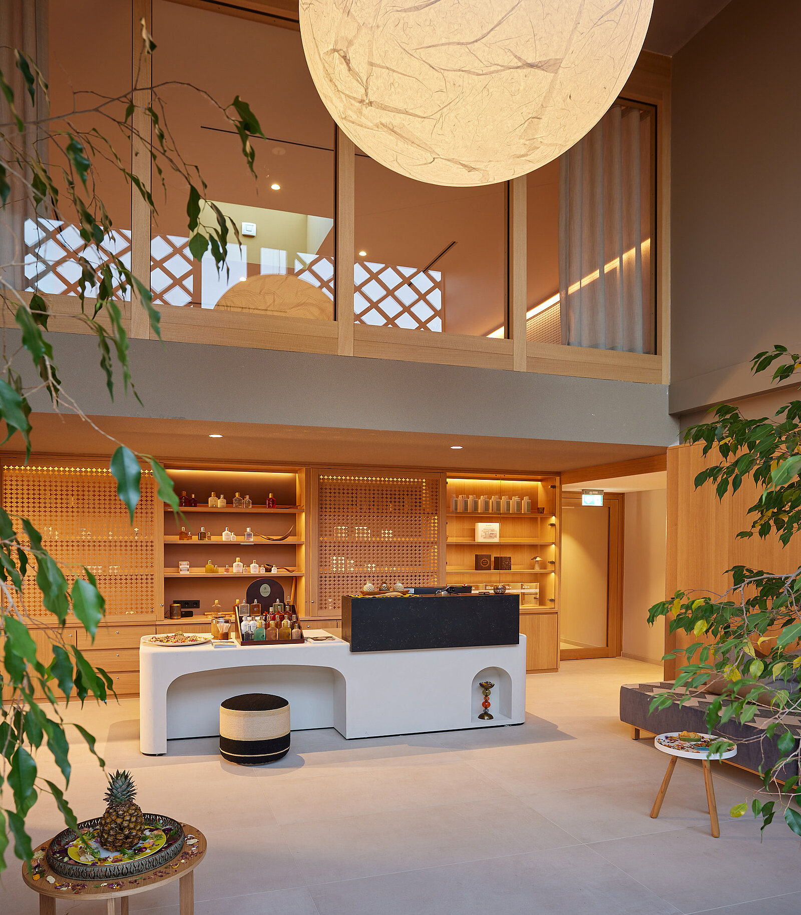 Ein moderner Empfangsbereich mit einem hellen und minimalistischen Design. Ein runder Leuchtkörper hängt von der Decke, während Regale mit Tee und anderen Produkten im Hintergrund sichtbar sind. Grüne Pflanzen bringen einen natürlichen Akzent in den Raum.