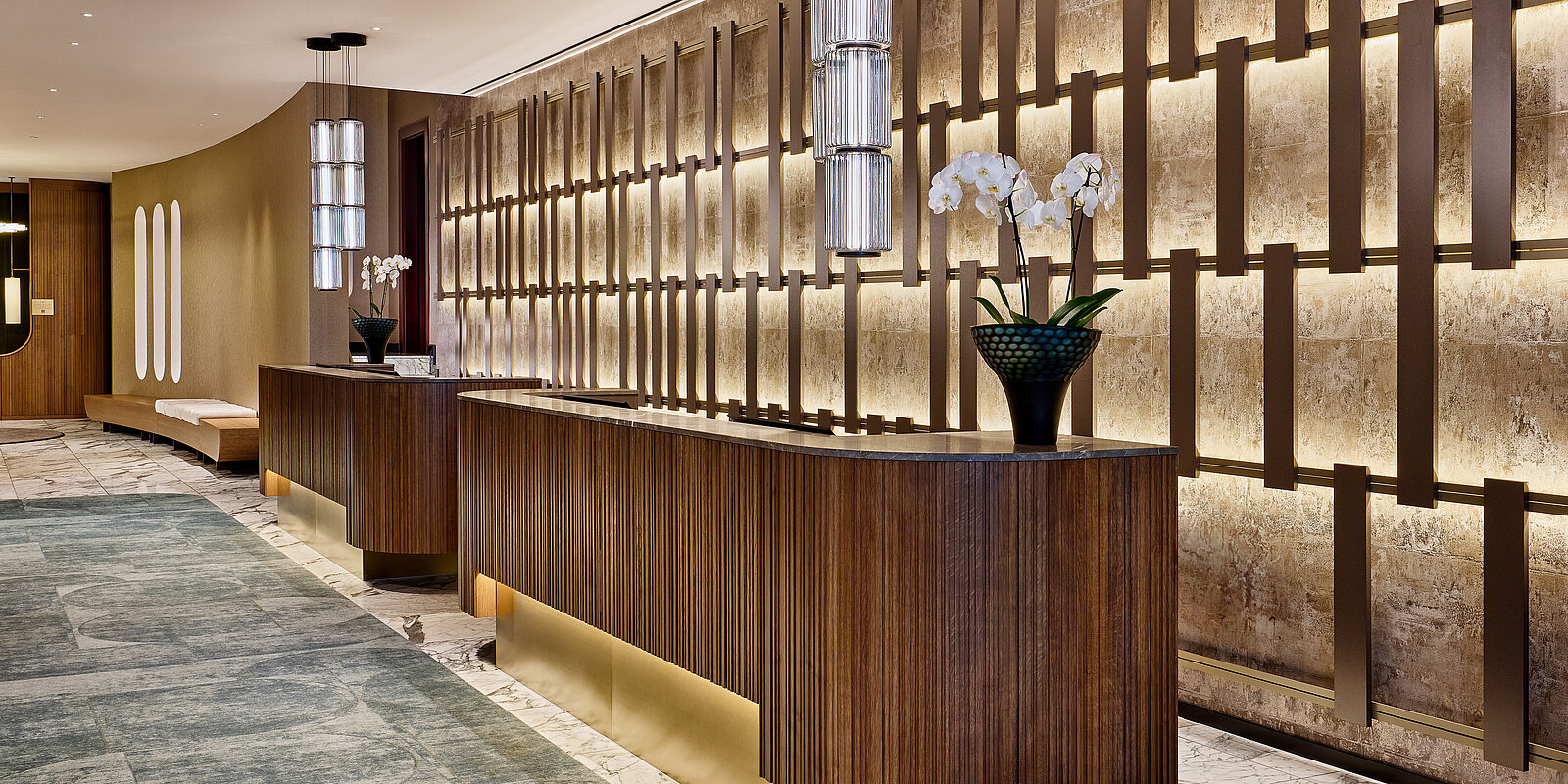 Ein eleganter Hotel-Lobby-Bereich mit einem langen, geschwungenen Empfangstresen aus Holz. An der Rückwand befinden sich Akzentbeleuchtungen und moderne Dekorationen, darunter Orchideen in einer stilvollen Vase. Die Atmosphäre wirkt einladend und luxuriös.