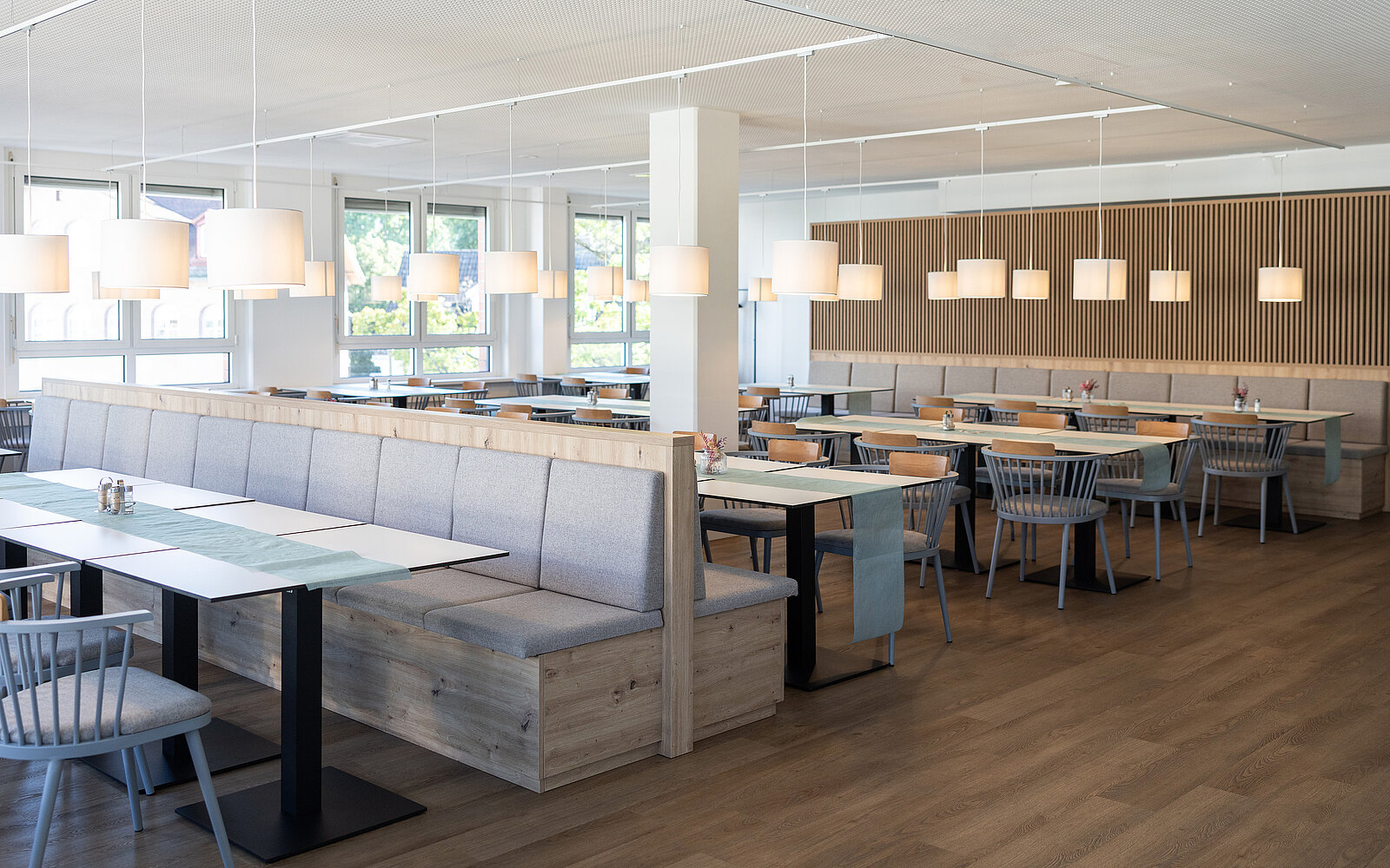 Ein modernes und helles Restaurant mit Holztischen und grauen Stühlen. Es gibt gepolsterte Sitzbänke und hängende Lampen. Große Fenster lassen viel Tageslicht herein. Der Raum wirkt einladend und geräumig.