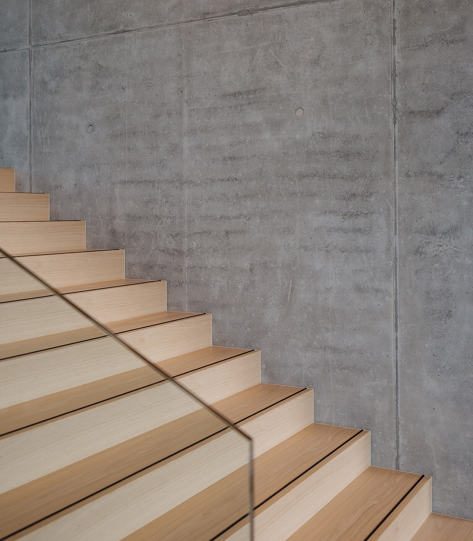 Nahaufnahme einer Holztreppe mit hellen Stufen, die sich durch klare Linien und ein elegantes Design auszeichnet. Die Treppe führt an einer glatten, grauen Betonwand empor und unterstreicht die moderne architektonische Ästhetik. Ein Glasgeländer sorgt für Halt an der Seite.