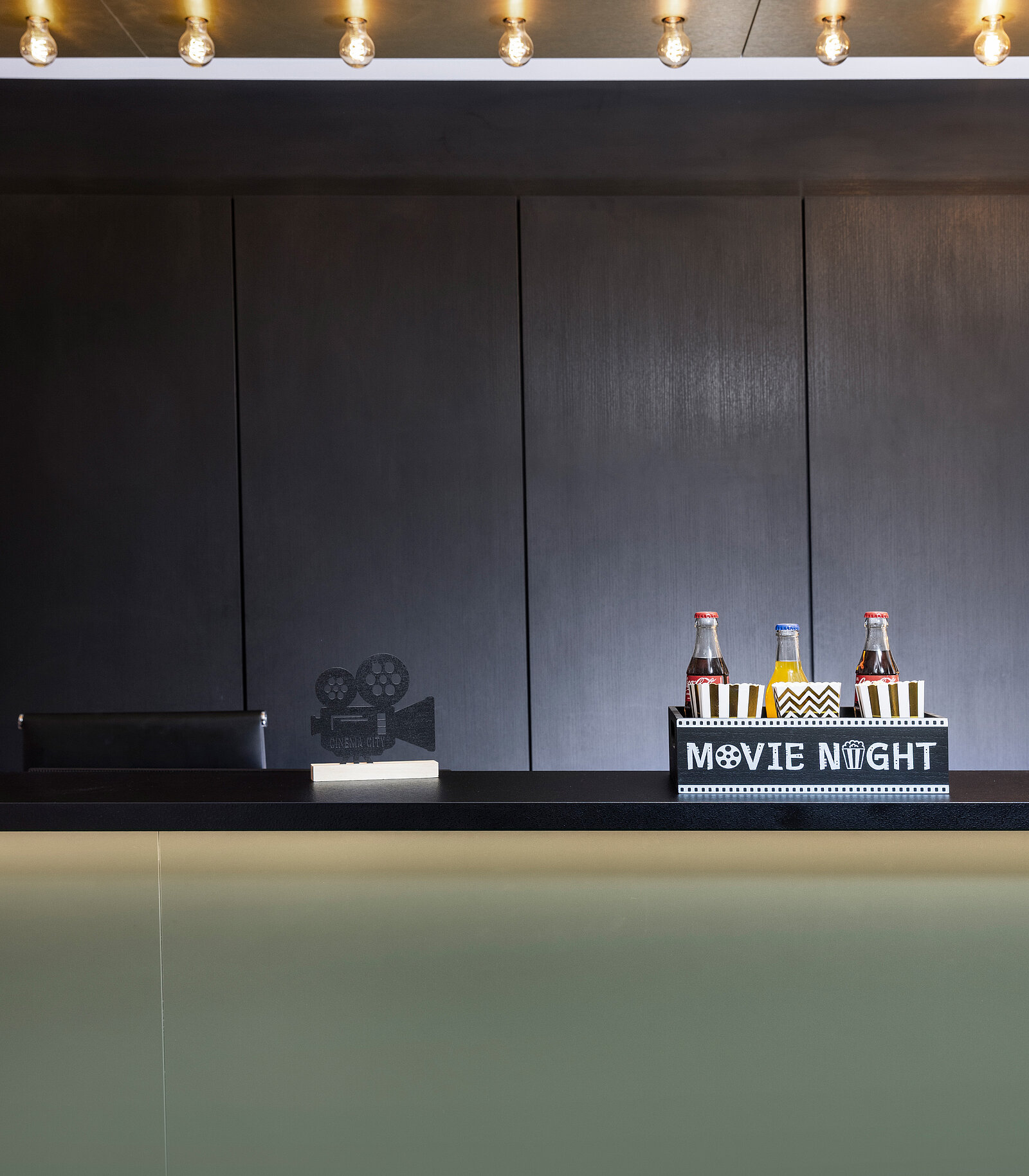 A dimly lit movie-themed display features a minimalistic counter with two beverage bottles and a striped background, labeled "MOVIE NIGHT." A decorative film reel silhouette adds to the cinematic ambiance, creating a cozy setting for a movie viewing experience.