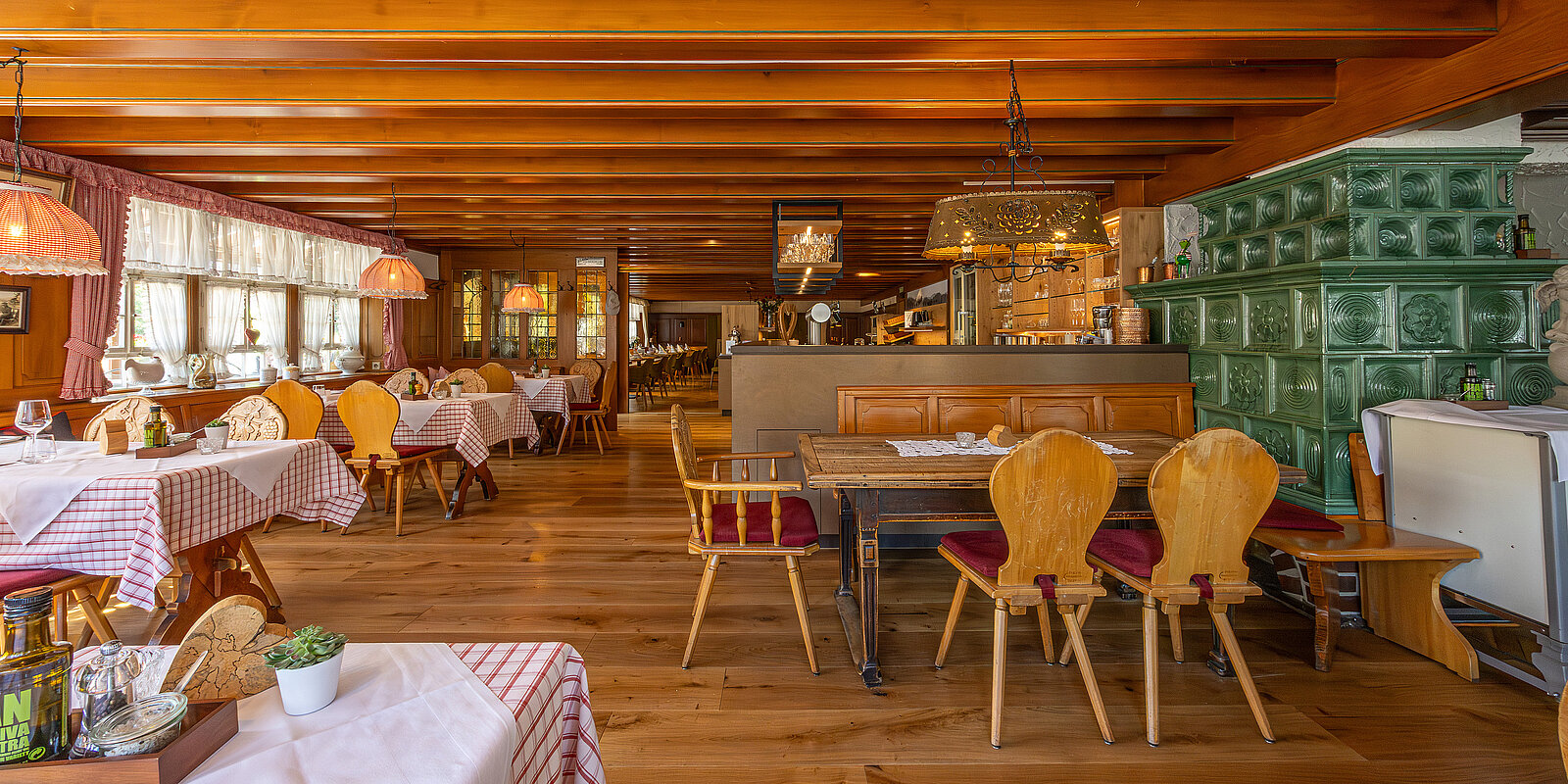 Gemütliches Restaurant im Landhausstil mit Holzmöbeln und karierten Tischdecken. Ein grüner Kachelofen steht im Raum. Die Atmosphäre ist warm und einladend, geeignet für entspannte Mahlzeiten. Große Fenster lassen viel Tageslicht hinein.