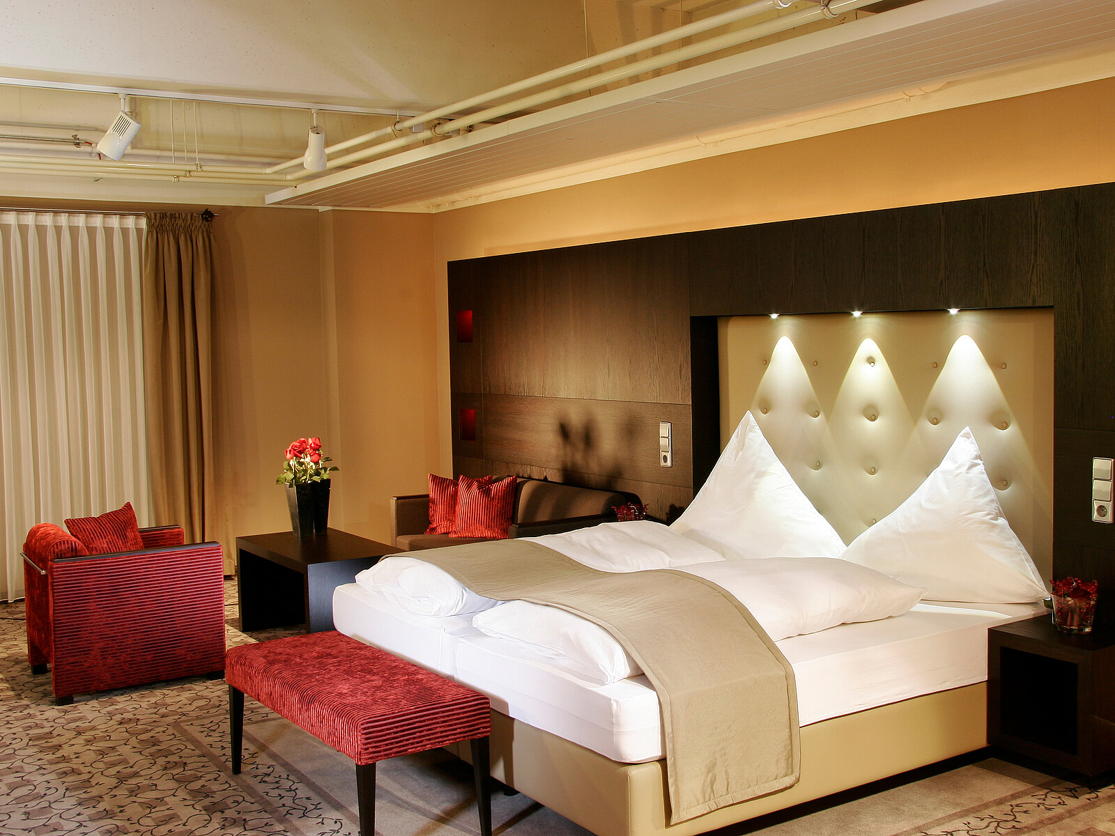 Ein modernes Schlafzimmer mit einem großen Doppelbett, das geperkt ist. Hinter dem Bett befindet sich eine geschmackvolle Wand mit Beleuchtung. Neben dem Bett steht ein eleganter Tisch und zwei rote Sessel. Vorhänge hängen an einem großen Fenster, und ein Blumenarrangement ergänzt die Dekoration.