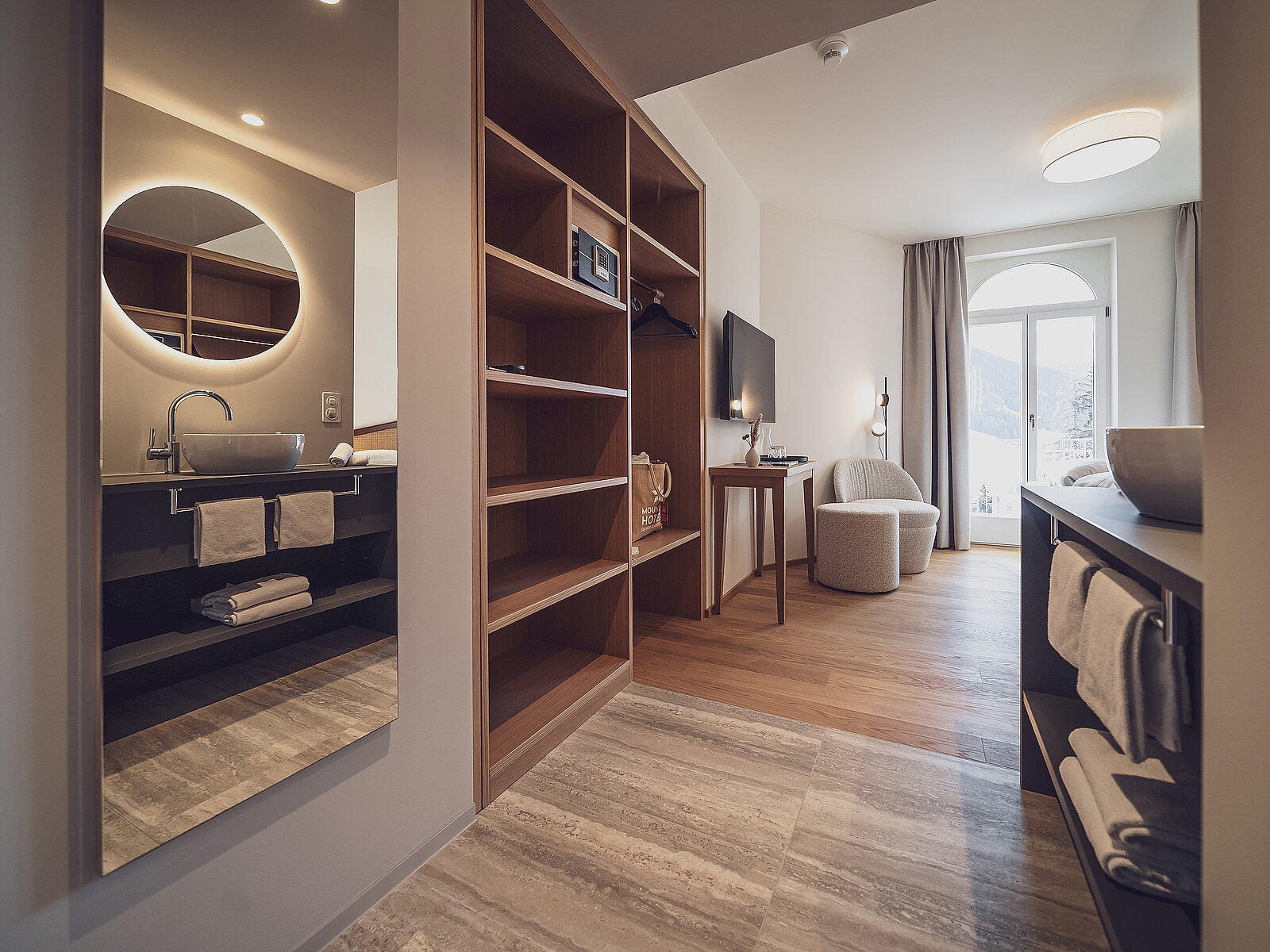 Modernes Hotelzimmer mit hellem Holzboden und großem Fenster. Im Vordergrund ist ein Badezimmer mit rundem Spiegel und Waschbecken sichtbar. Im Zimmer stehen ein Sessel und ein Tisch, daneben ein Fernseher. Elegantes, schlichtes Design mit einem hellen, entspannten Ambiente.