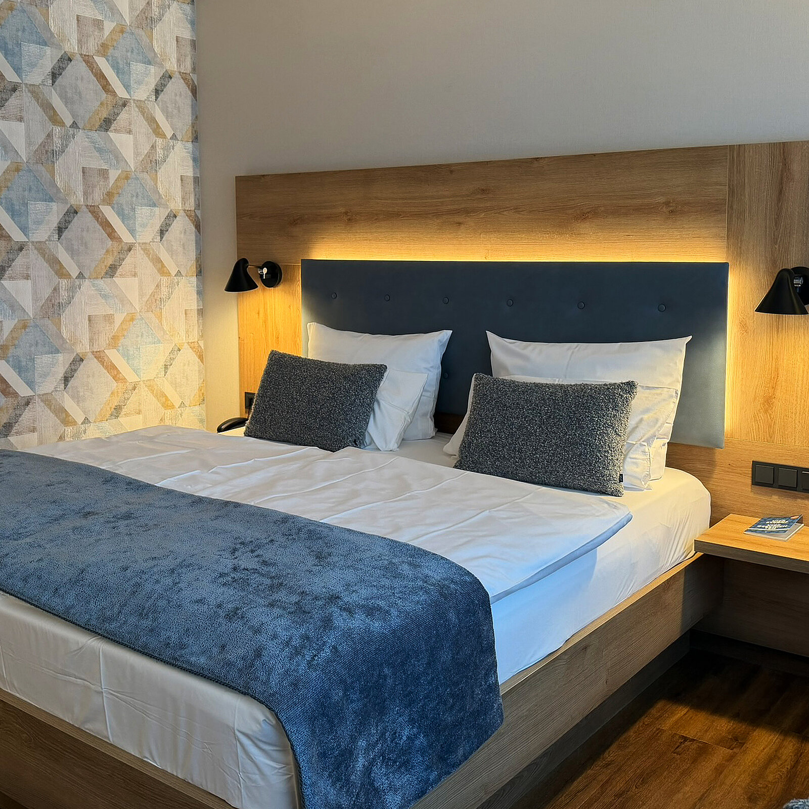 Ein modernes Schlafzimmer mit einem großen Bett, das mit weißen Laken und dekorativen Kissen ausgestattet ist. An der Wand hinter dem Bett befindet sich eine sanfte Beleuchtung, und neben dem Bett steht ein Nachttisch. Die Wände sind mit einem geometrischen Muster dekoriert.
