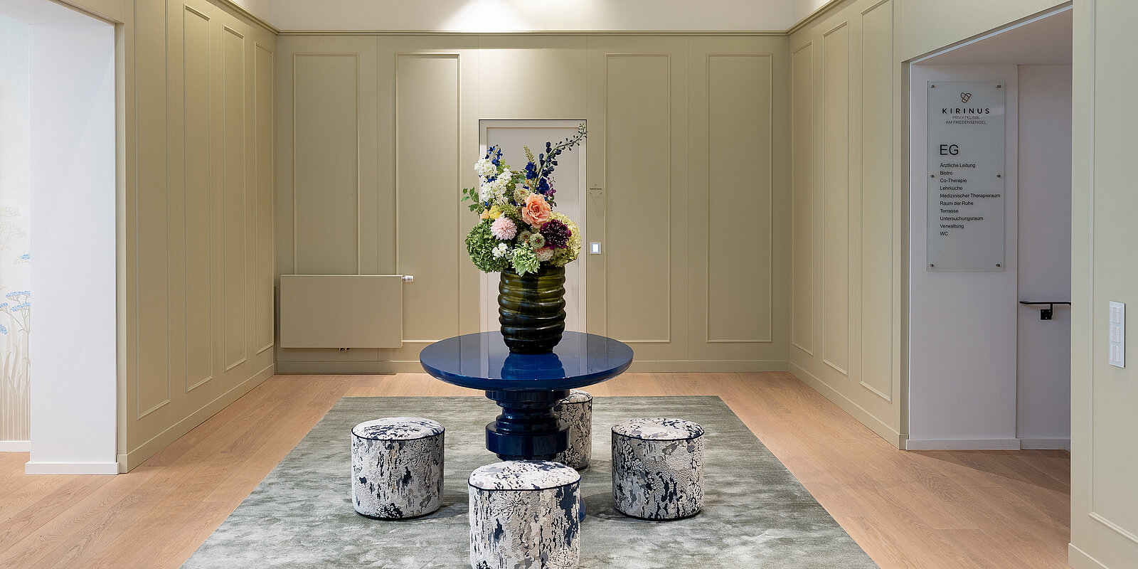Ein eleganter Raum mit hellem Holzboden zeigt in der Mitte einen blauen Tisch mit einem Blumenarrangement. Vier gepolsterte Hocker umgeben den Tisch auf einem grauen Teppich. Die Wände sind in einem hellen Beigeton gehalten.