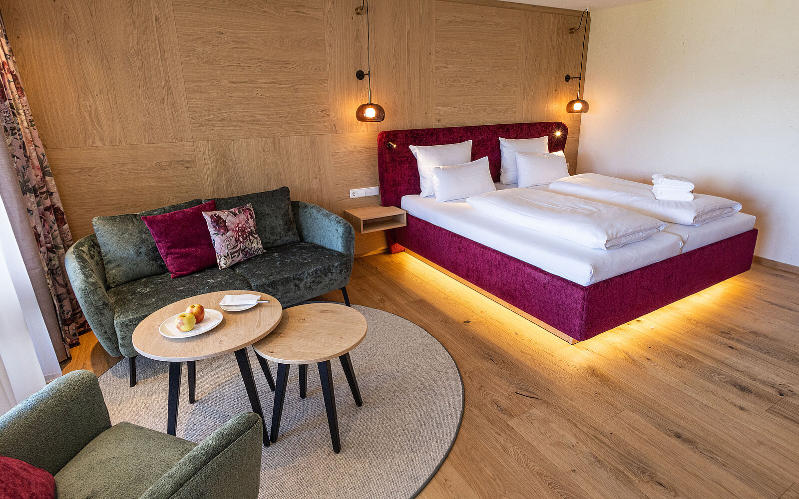Gemütliches Hotelzimmer mit einem großen Bett, beleuchtetem Sockel und zwei weißen Kissen. Links steht ein grünes Sofa neben einem runden Teppich und zwei Holztischen, auf denen Obst liegt. Die Wände aus hellem Holz und die warmen Farben schaffen eine einladende Atmosphäre.
