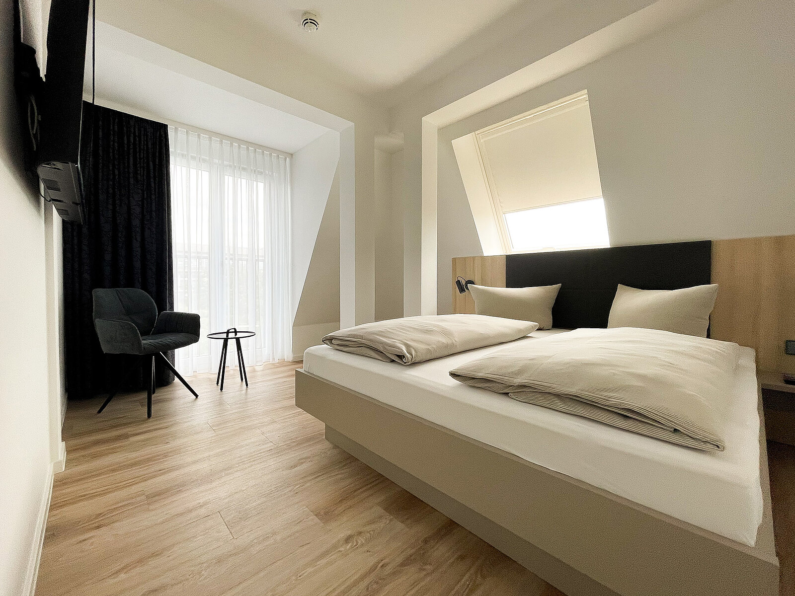 Ein modernes Hotelzimmer mit einem großen Bett, zwei Kissen und einer hellen Bettdecke. Im Hintergrund befindet sich ein Sessel neben einem kleinen runden Tisch. Tageslicht strömt durch ein großes Fenster mit einem dunklen Vorhang. Der Boden ist aus Holz.