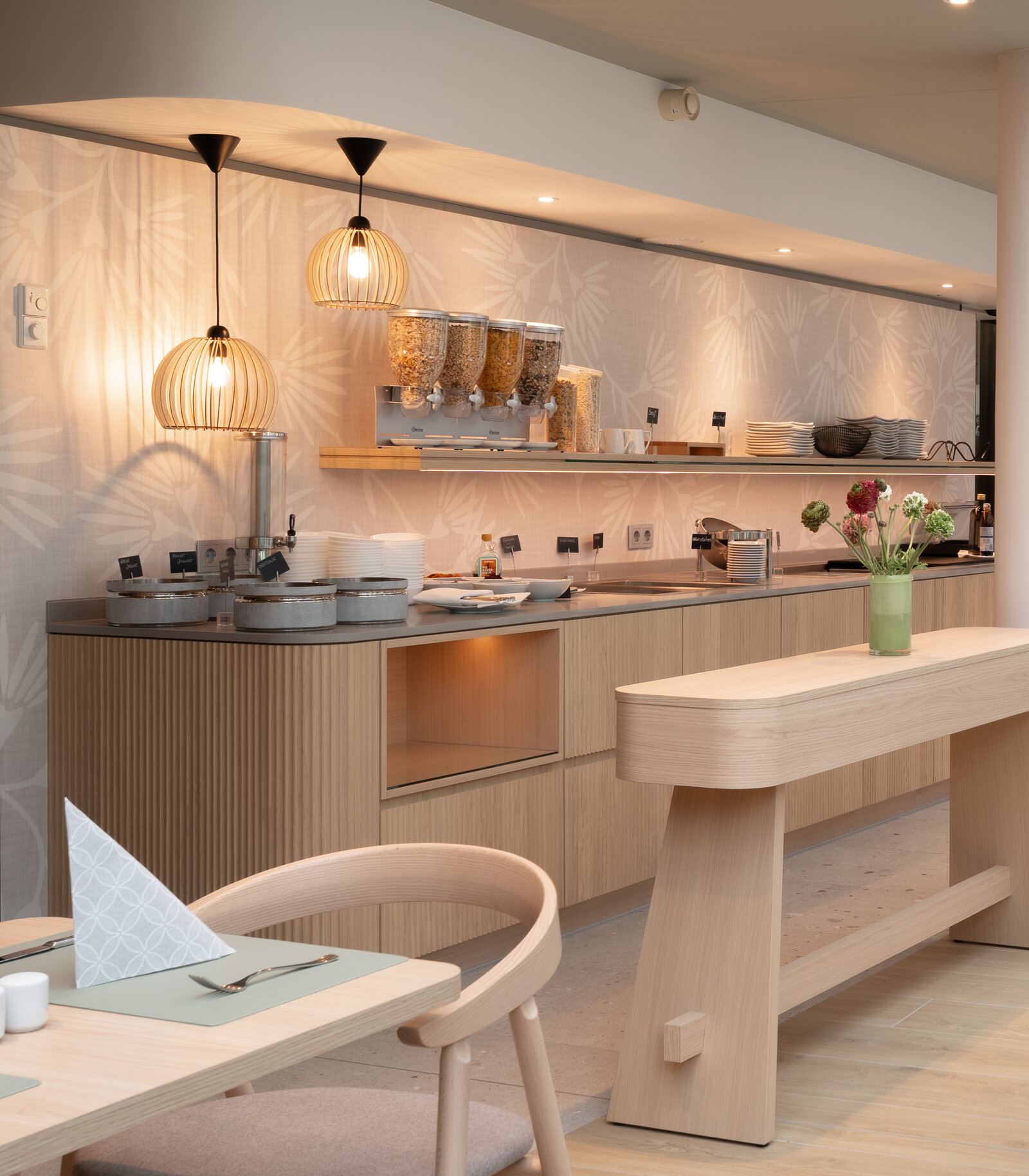 Modern breakfast buffet with cereal dispensers, hanging lamps, and neatly arranged dishes. Light wood furniture and a vase with flowers on the table.
