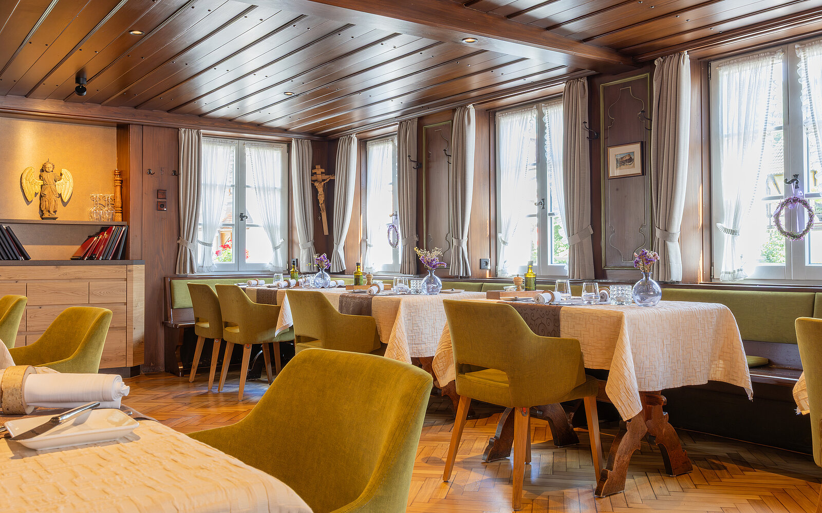 Gemütliches Restaurant mit Holzmöbeln und -decke. Tische mit weißen Tischdecken sind dekoriert mit Blumenvasen. Große Fenster lassen viel Tageslicht herein. Grün gepolsterte Stühle umgeben die Tische, und im Hintergrund sind Regale zu sehen.