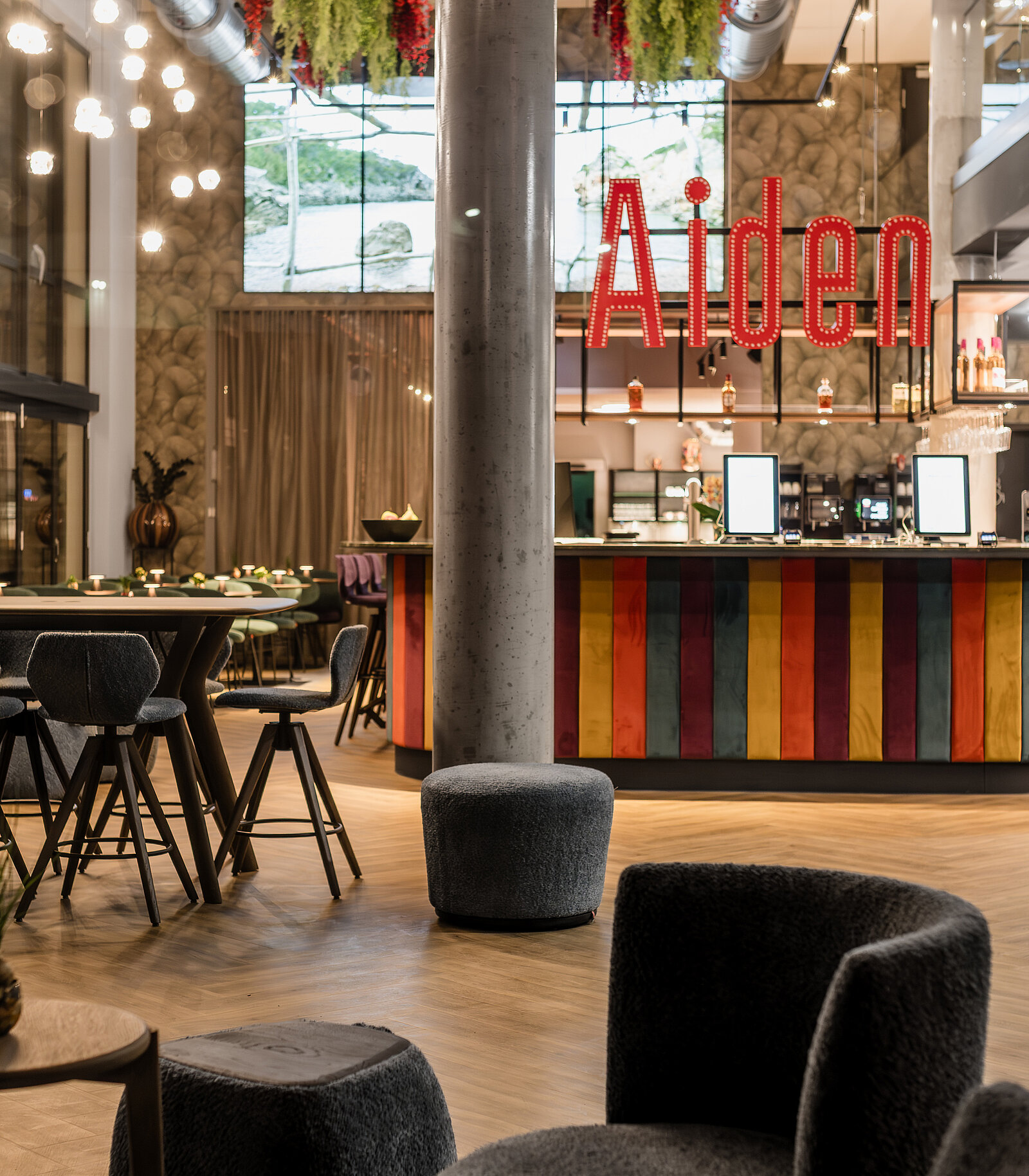 Gemütliches Café mit moderner Einrichtung. Bunte Bar mit dem Schriftzug „Aiden“ in roten Leuchtbuchstaben. Hohe runde Tische und Barhocker, warme Beleuchtung. Pflanzen und große Fenster im Hintergrund sorgen für ein einladendes Ambiente.
