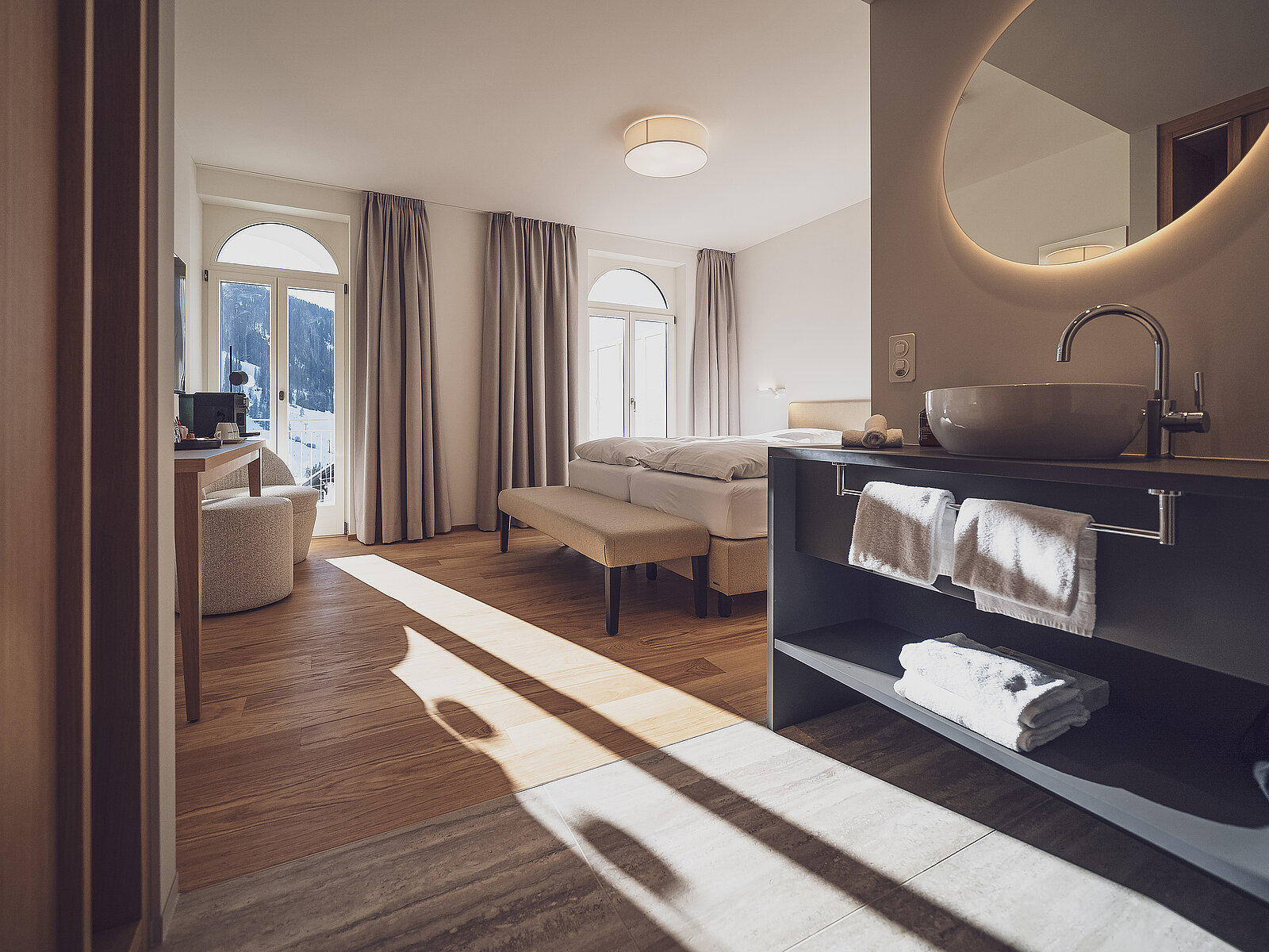 Helles und elegantes Hotelzimmer mit großem Bett, bodentiefen Fenstern und Balkonblick. Im Vordergrund ein modernes Waschbecken mit Handtüchern. Warme Holzböden und weiche Beleuchtung schaffen eine einladende Atmosphäre.