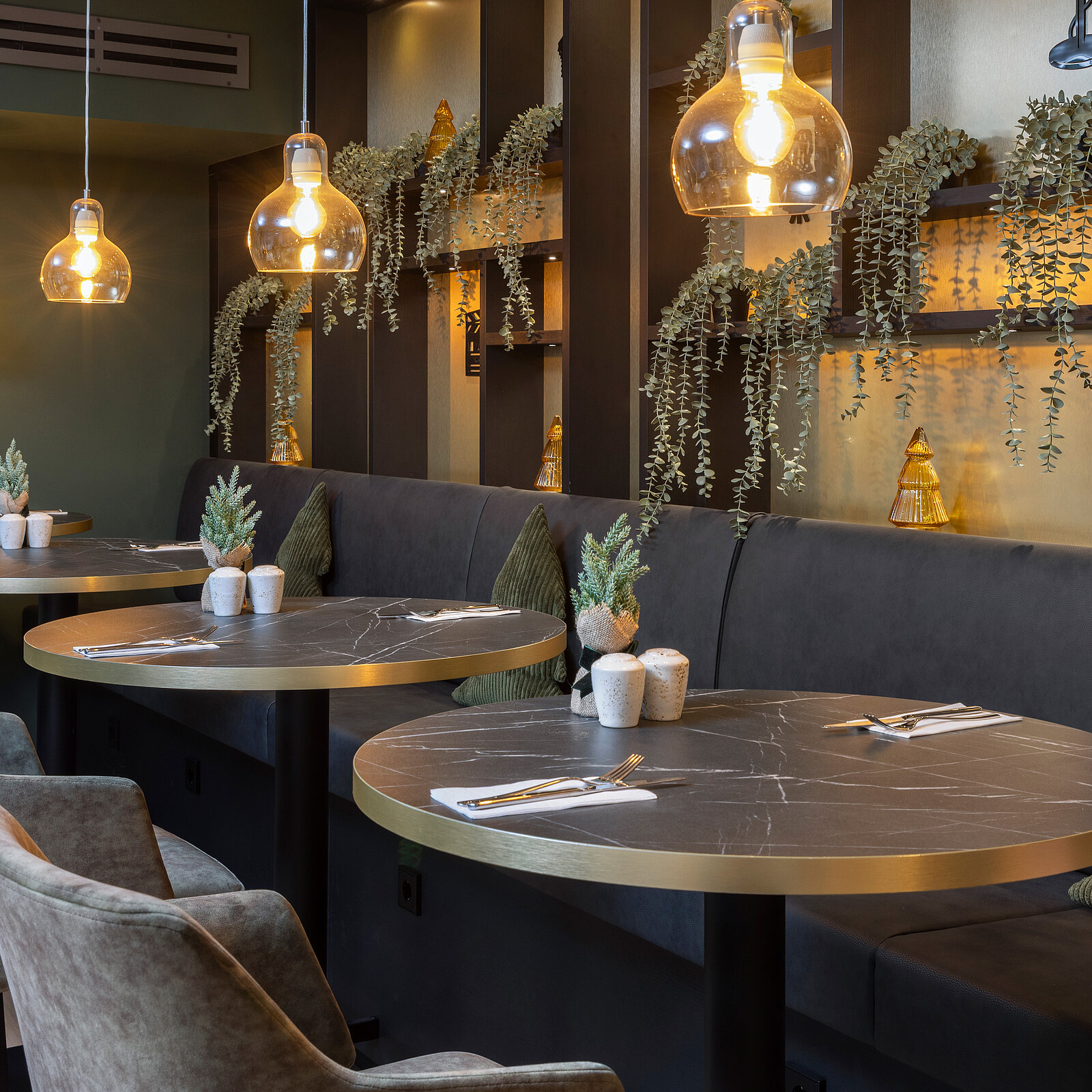 A stylish restaurant interior featuring round tables with decorative plants at each setting. Soft lighting from hanging pendant lamps illuminates the space, complemented by dark walls and green plants. Cozy seating areas are arranged against a backdrop of shelves and greenery, creating an inviting atmosphere.