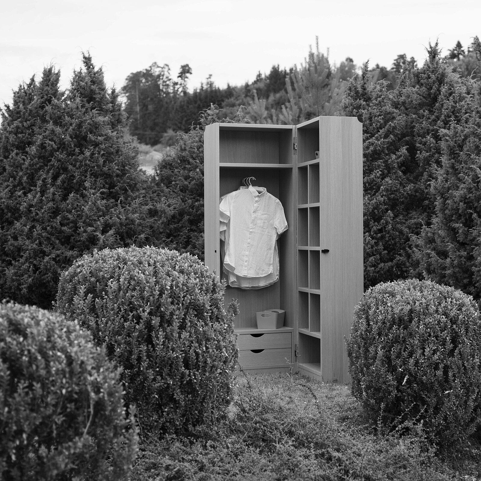 Ein offener Kleiderschrank steht zwischen runden, grünen Sträuchern. Im Inneren hängt ein weißes Hemd. Die Szene ist in Schwarzweiß gehalten und vermittelt eine melancholische Stimmung, die die Abgeschiedenheit und den Kontrast zwischen dem Kleiderschrank und der umgebenden Natur betont.