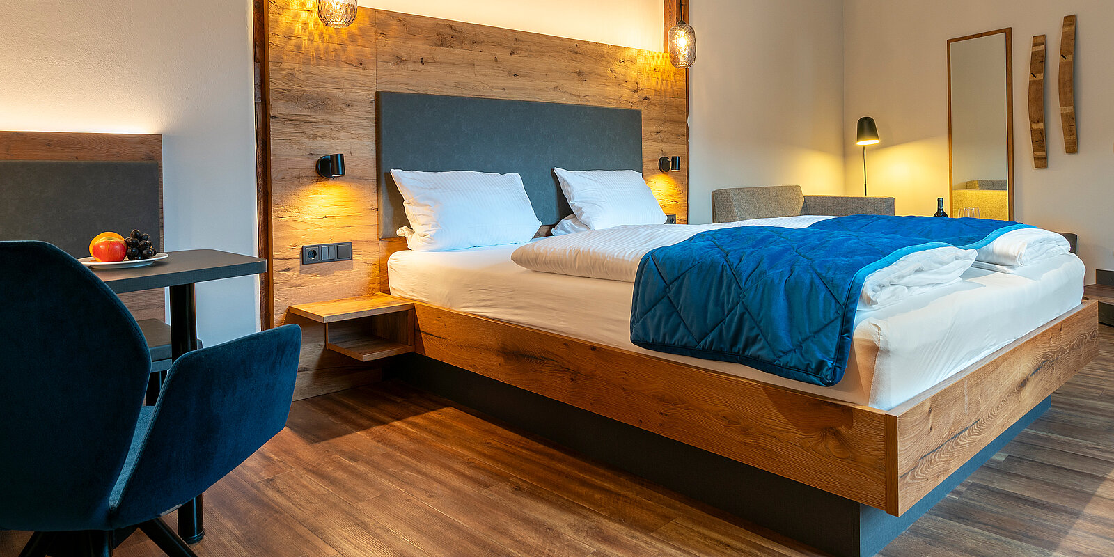 Ein einladendes Schlafzimmer mit einem großen, bequemen Bett, das mit weißen Kissen und einer blauen Bettdecke dekoriert ist. Daneben steht ein kleiner Tisch mit einem Stuhl. Der Raum hat eine warme Holzoptik und sanfte Beleuchtung.