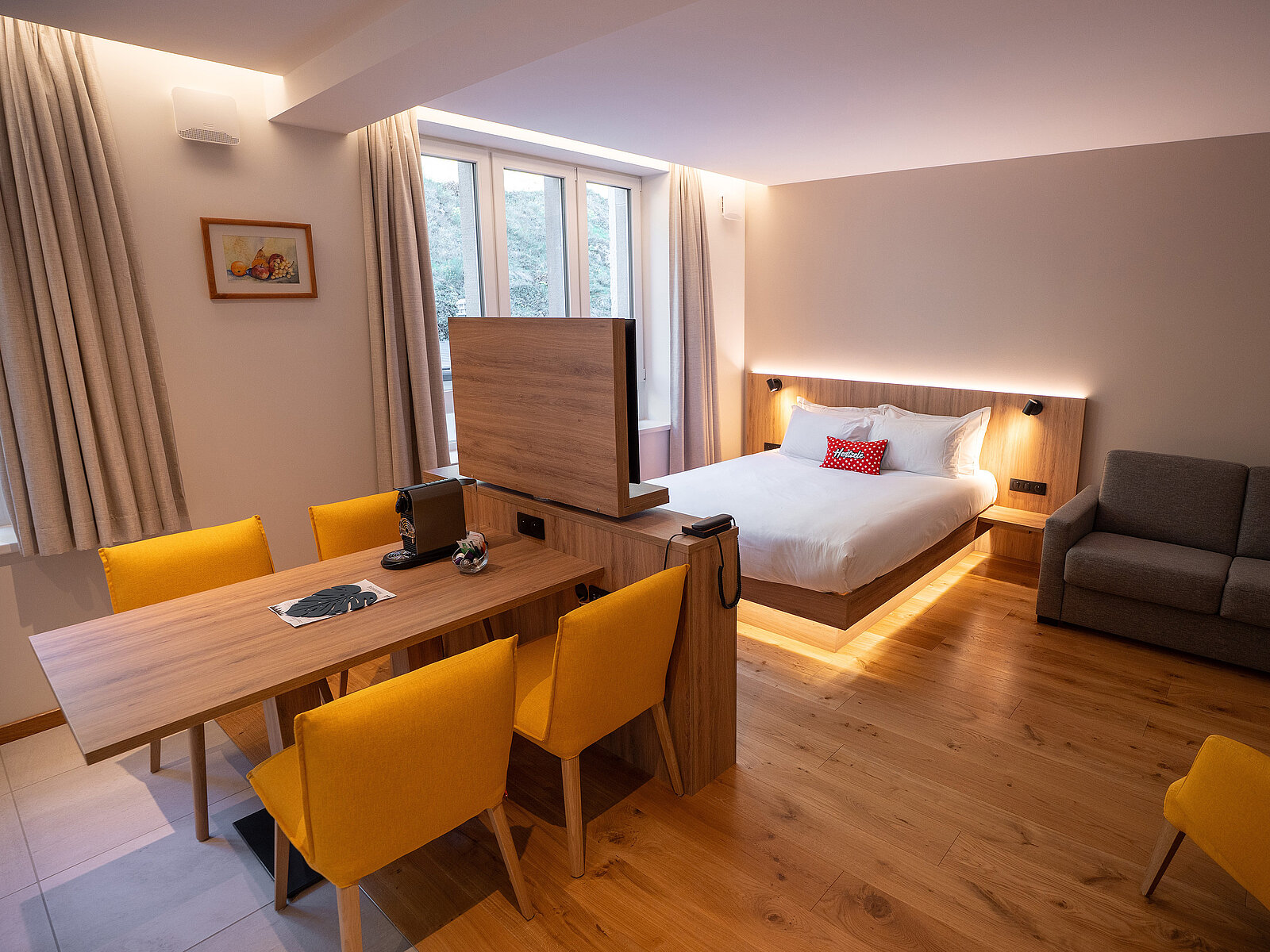 Ein modernes Hotelzimmer mit einem Doppelbett, einem Sofa, einem Holztisch und gelben Stühlen. Der Raum ist hell erleuchtet und verfügt über Holzfußboden sowie große Fenster mit Vorhängen. Eine dezente Beleuchtung strahlt hinter dem Bett hervor.