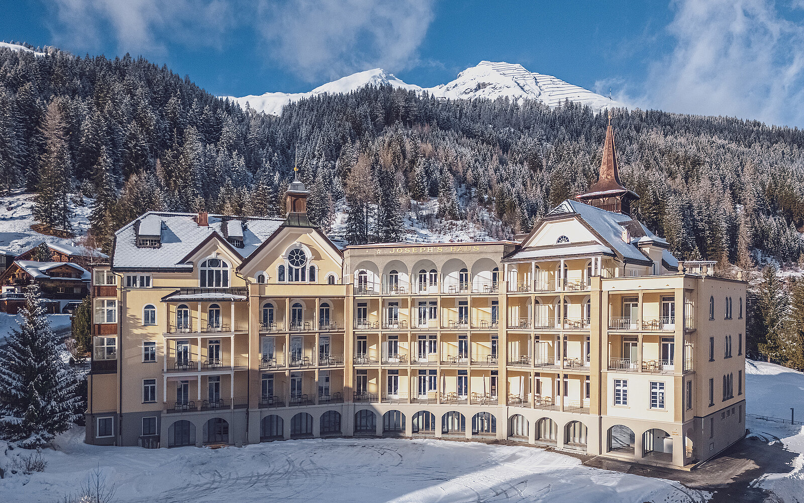 Ein großes, elegantes Hotelgebäude im alpinen Stil steht vor einer schneebedeckten Bergkulisse. Die Wände sind hell, und das Dach ist mit Schnee bedeckt. Im Hintergrund sind bewaldete Berghänge und verschneite Gipfel unter einem blauen Himmel sichtbar.