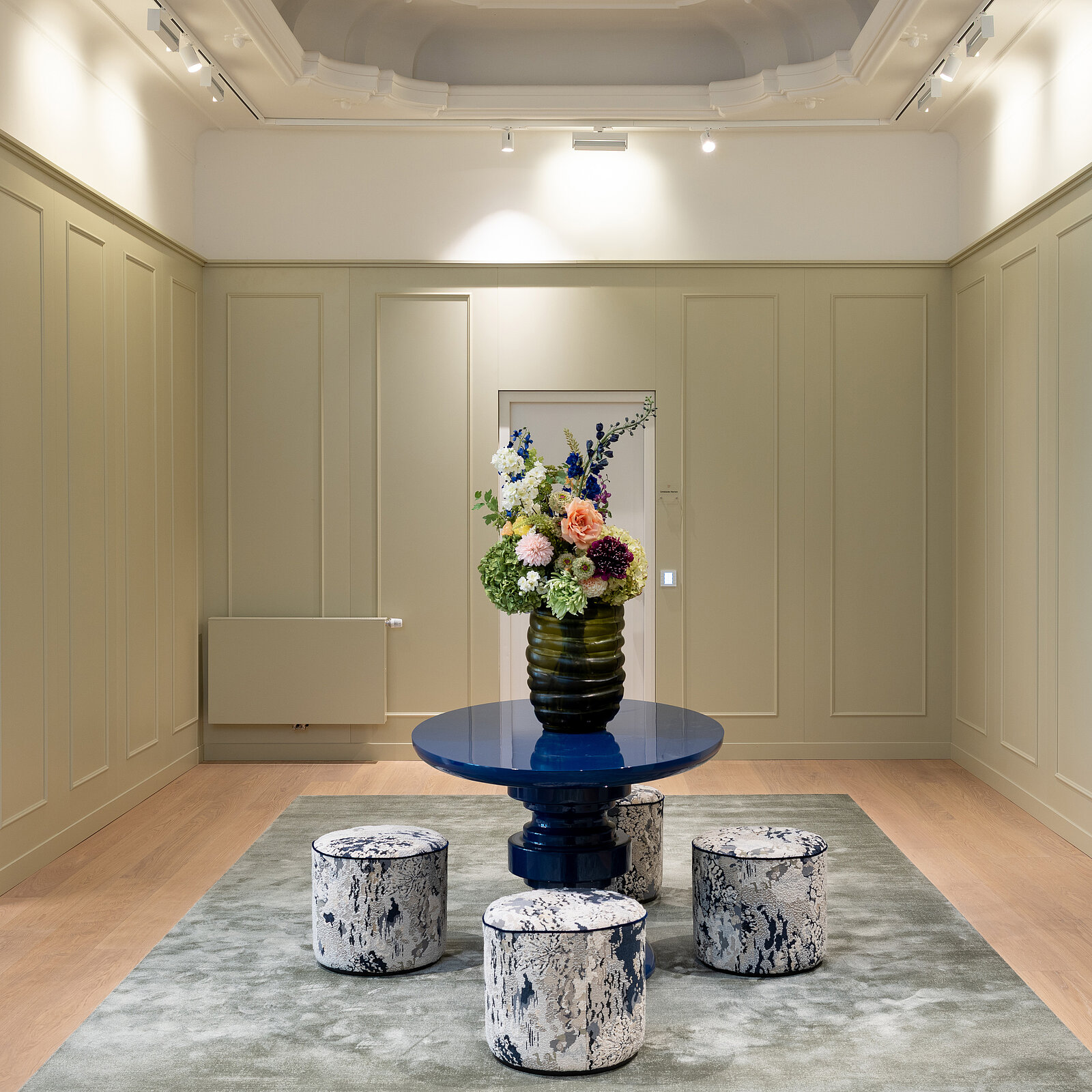 Ein eleganter Raum mit einem runden blauen Tisch in der Mitte, darauf eine Vase mit bunten Blumen. Drei Hocker mit marmornem Muster stehen um den Tisch. Der Boden ist mit einem grauen Teppich bedeckt.