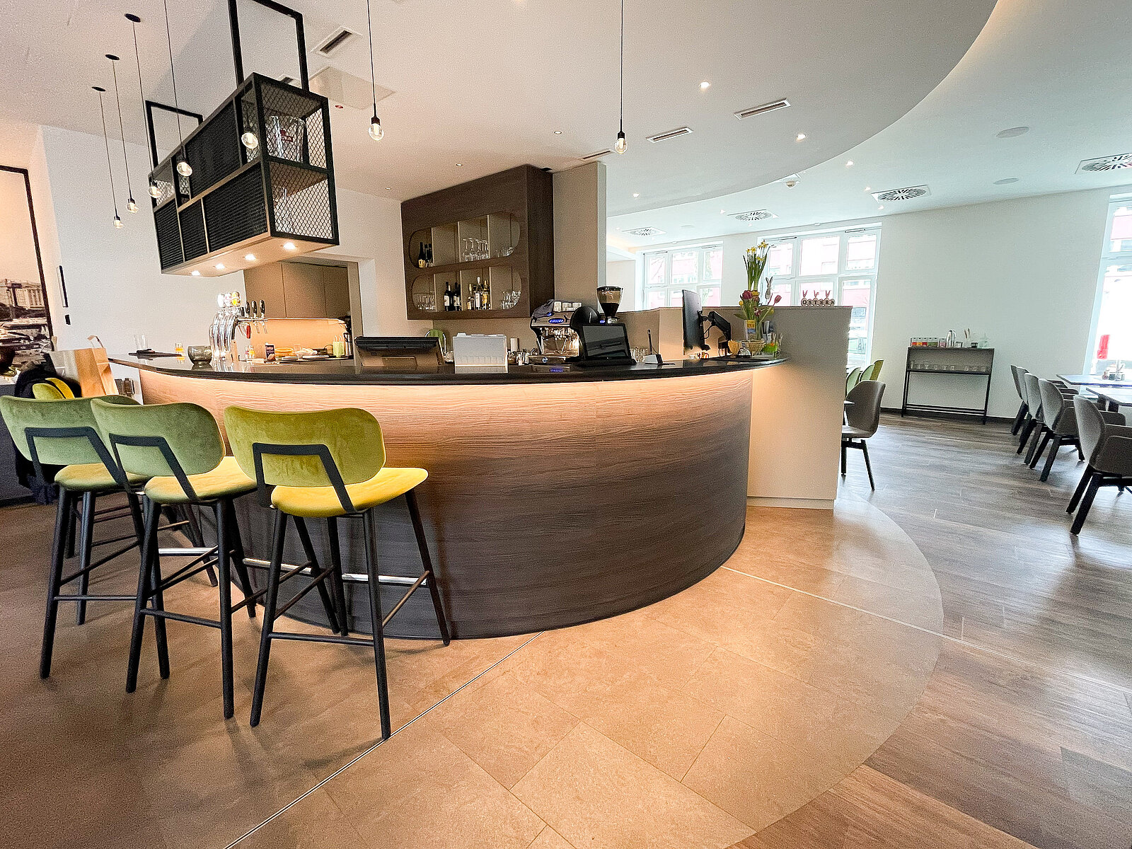Moderne, helle Café-Bar mit geschwungenem Tresen, drei hohen, gelben Barhockern und eleganten Hängeleuchten. Im Hintergrund sind Regale mit Gläsern und eine Kaffeemaschine zu sehen. Rechts steht ein kleiner Tisch mit Vase und Blumen nahe der Fensterfront.