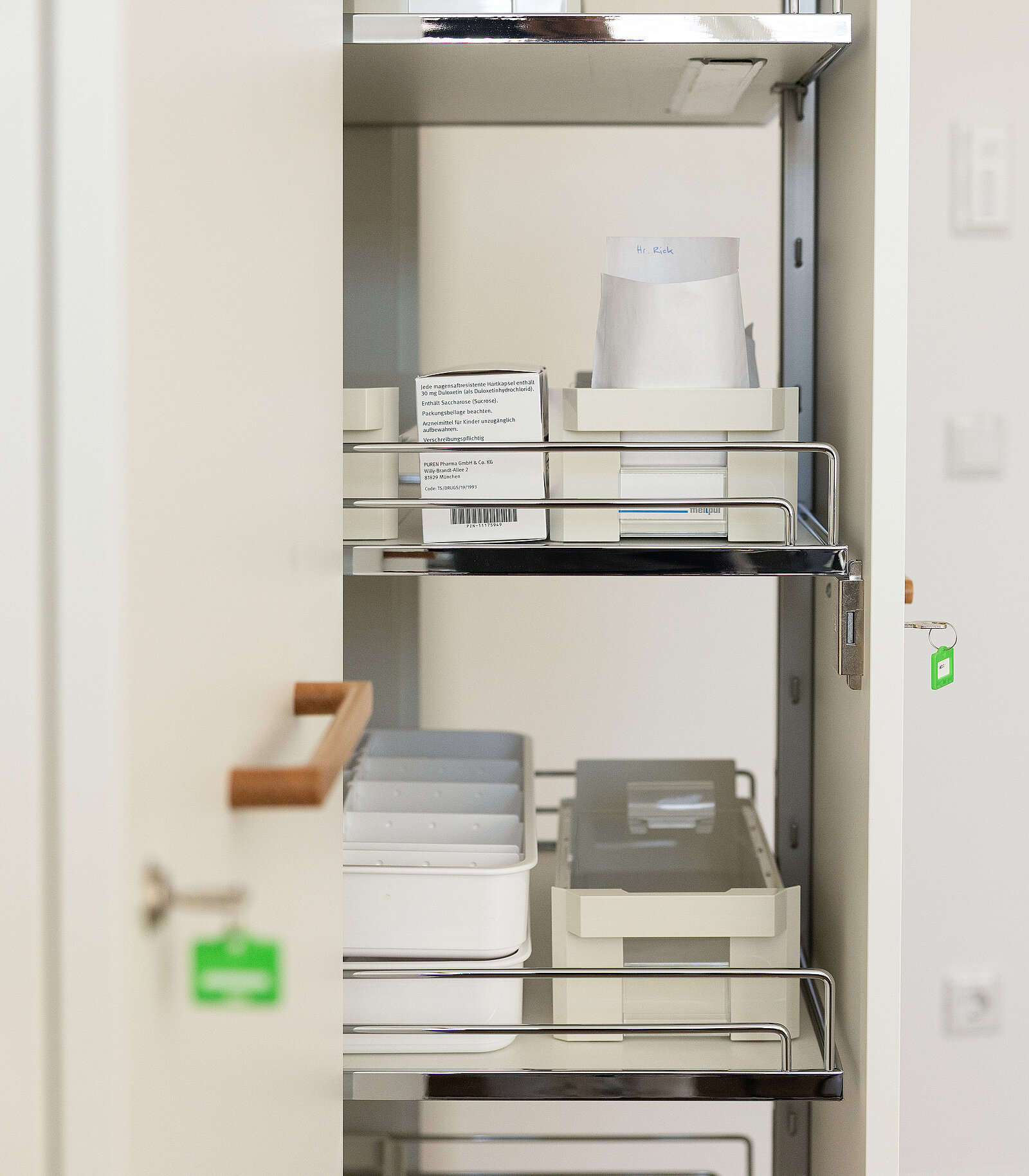 Ein geöffneter Medizinschrank zeigt mehrere Regale mit Tabletts, die Papierdokumente und andere medizinische Utensilien enthalten. Die Tür des Schranks ist teilweise geöffnet.