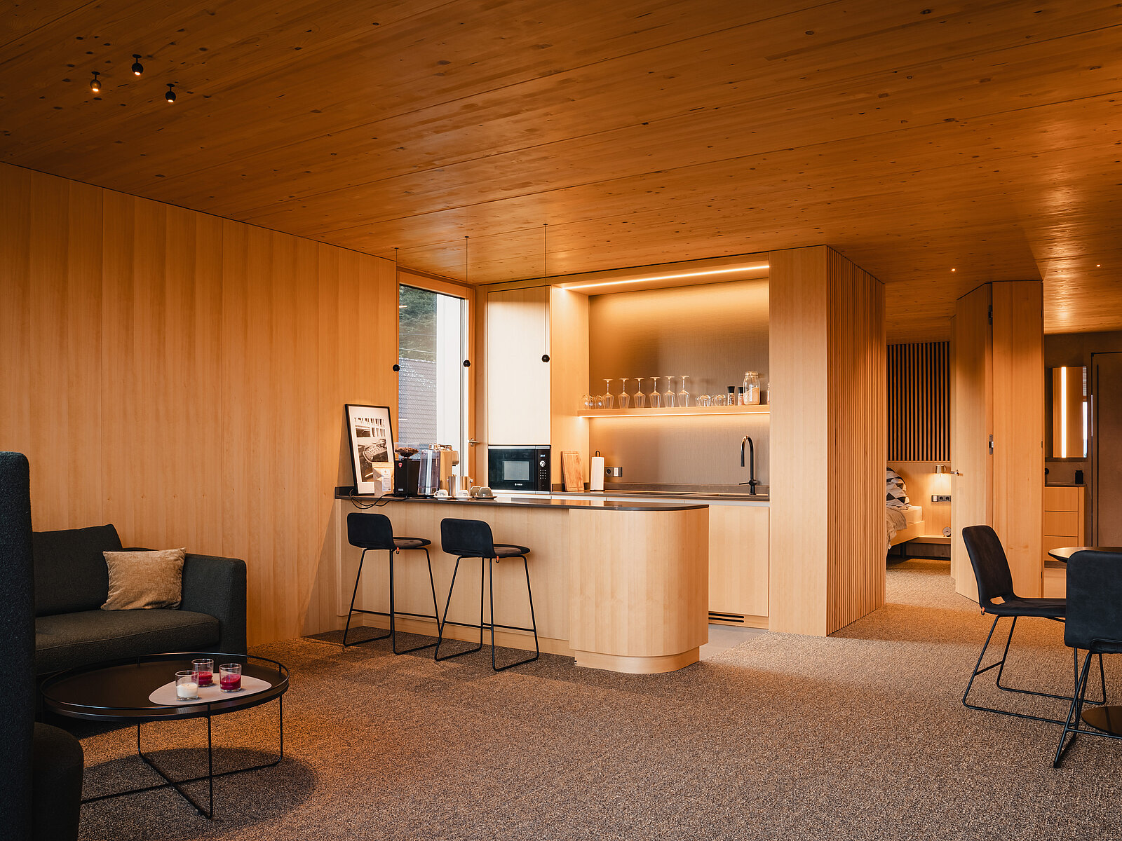 Ein eleganter, minimalistischer Wohnraum mit Holzwänden und -decken. Eine moderne Küchenzeile mit Barhockern ist sichtbar. Der Raum wirkt warm und einladend, ausgestattet mit stilvollen, dunklen Möbeln und warmem Licht. Ein Schlafzimmer ist im Hintergrund erkennbar.