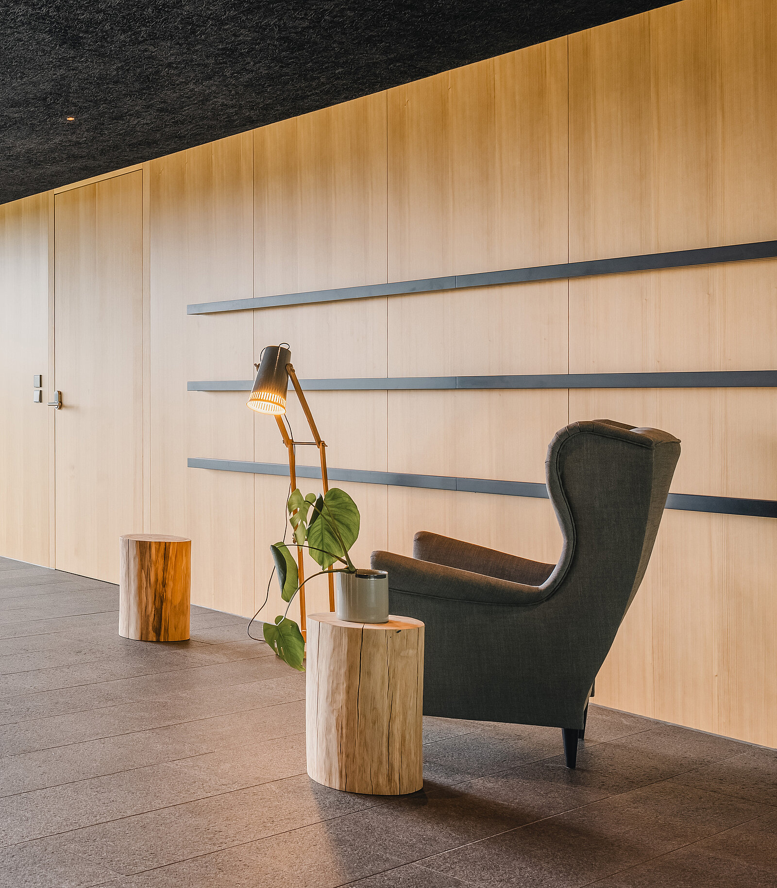 Ein stilvolles Interieur mit einem dunkelgrauen Sessel, einer Stehlampe und einem kleinen Pflanzenständer auf einer Holzstamm-Tischplatte. Der Hintergrund besteht aus hellen Holzwänden mit horizontalen Regalböden.