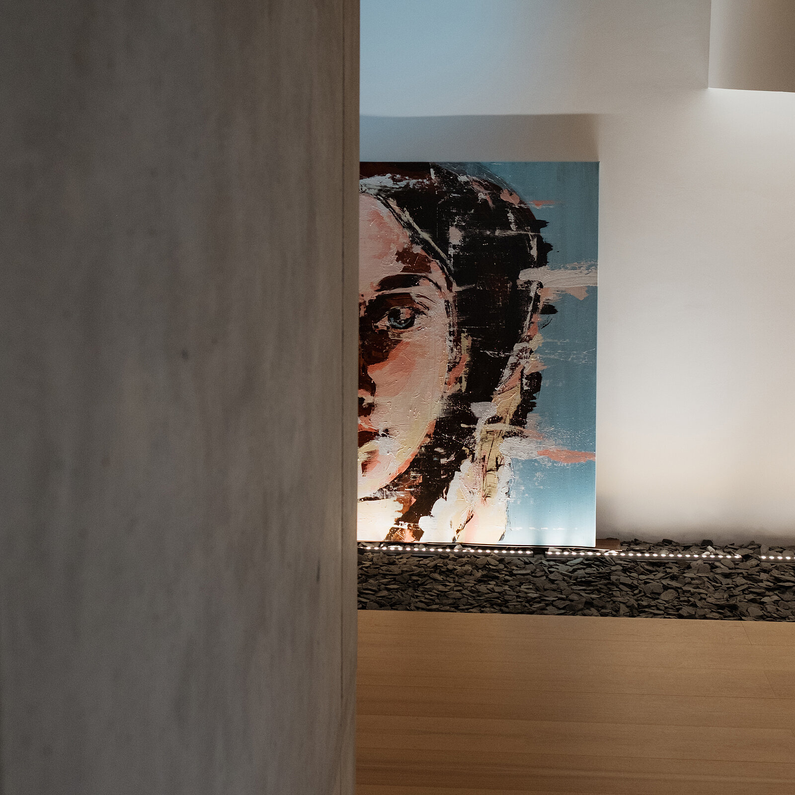 Das Bild zeigt ein Kunstwerk, das das Gesicht einer Frau mit einem nachdenklichen Ausdruck darstellt. Die Farben sind lebendig und kontrastreich, und der Hintergrund ist schlichte Architektur, die die Aufmerksamkeit auf die Frau lenkt.