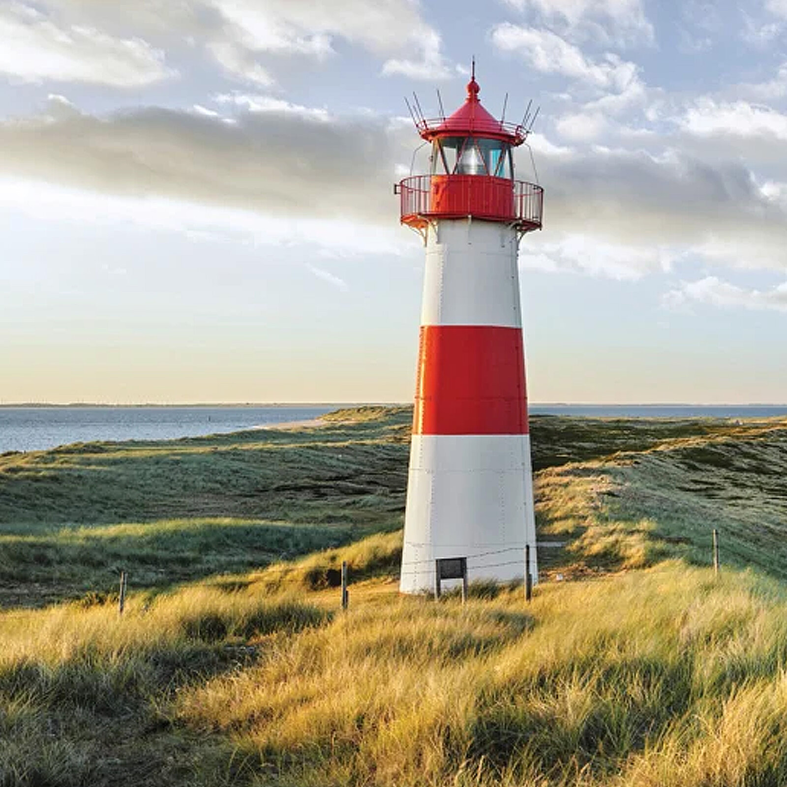 Ein rot-weiß gestreifter Leuchtturm steht inmitten von grünen Wiesen und sanften Hügeln. Im Hintergrund ist das Meer mit leicht bewölktem Himmel zu sehen.