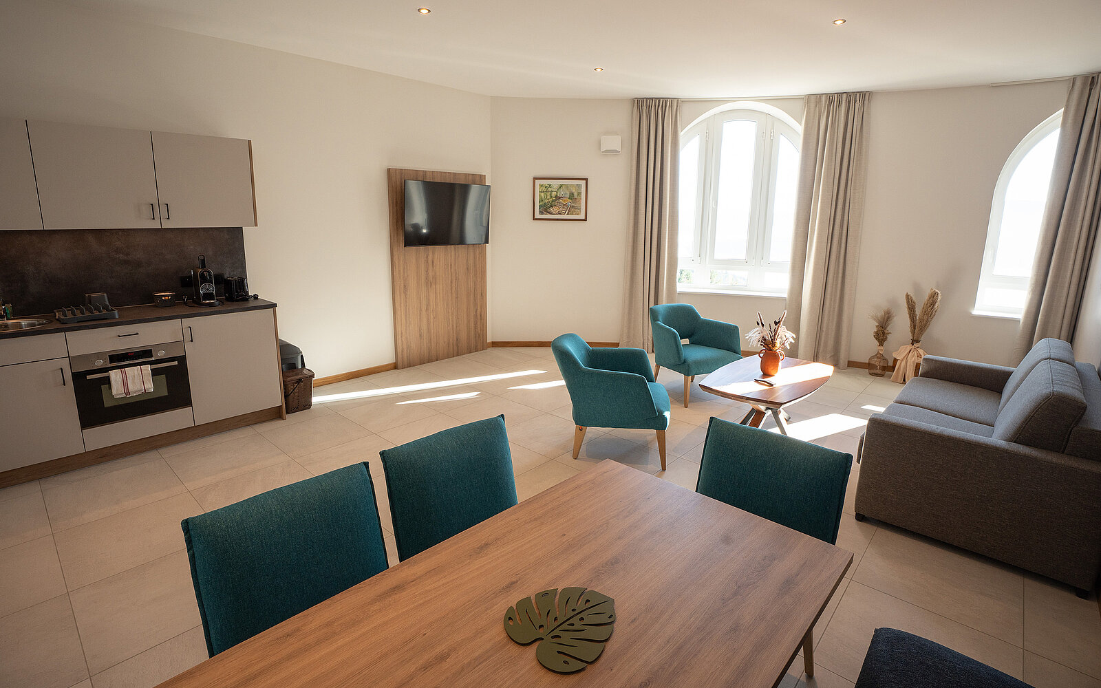 Eine moderne, helle Wohnküche mit Esstisch, Fernseher, Küchenzeile und gemütlichem Wohnbereich. Große Fenster lassen viel Licht herein.