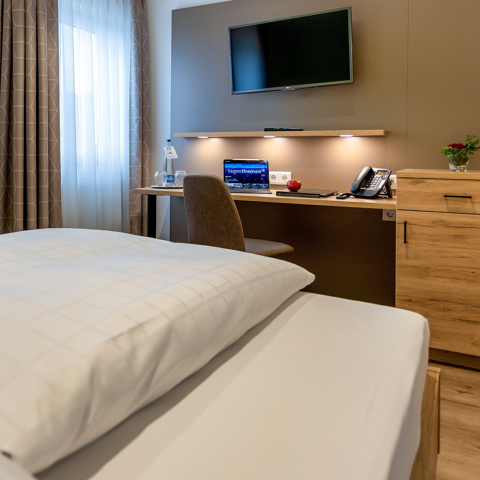 Ein modernes Hotelzimmer mit einem großen Bett, einem Schreibtisch, Fernseher an der Wand und einer Sitzgelegenheit. Auf dem Schreibtisch stehen eine Wasserflasche, ein Glas und ein Apfel. Rechts daneben befindet sich ein Holzschrank.