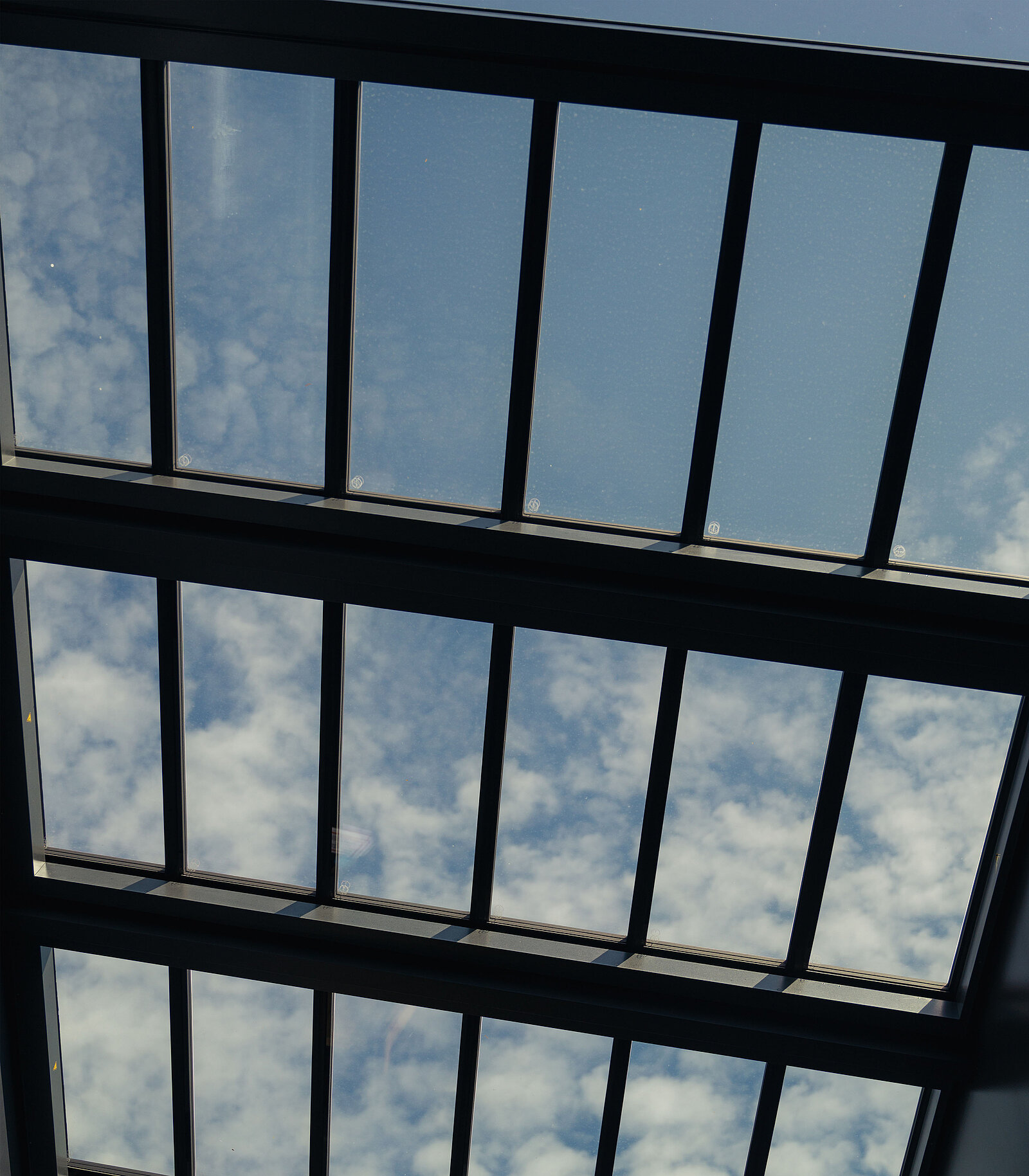 Eine Ansicht nach oben zu einem Oberlicht mit mehreren Glasfeldern. Die Scheiben sind schwarz gerahmt und geben den Blick auf den blauen Himmel frei, durch den weiche, weiße Wolken hindurchscheinen. Das Licht ist hell und schafft eine luftige, offene Atmosphäre.
