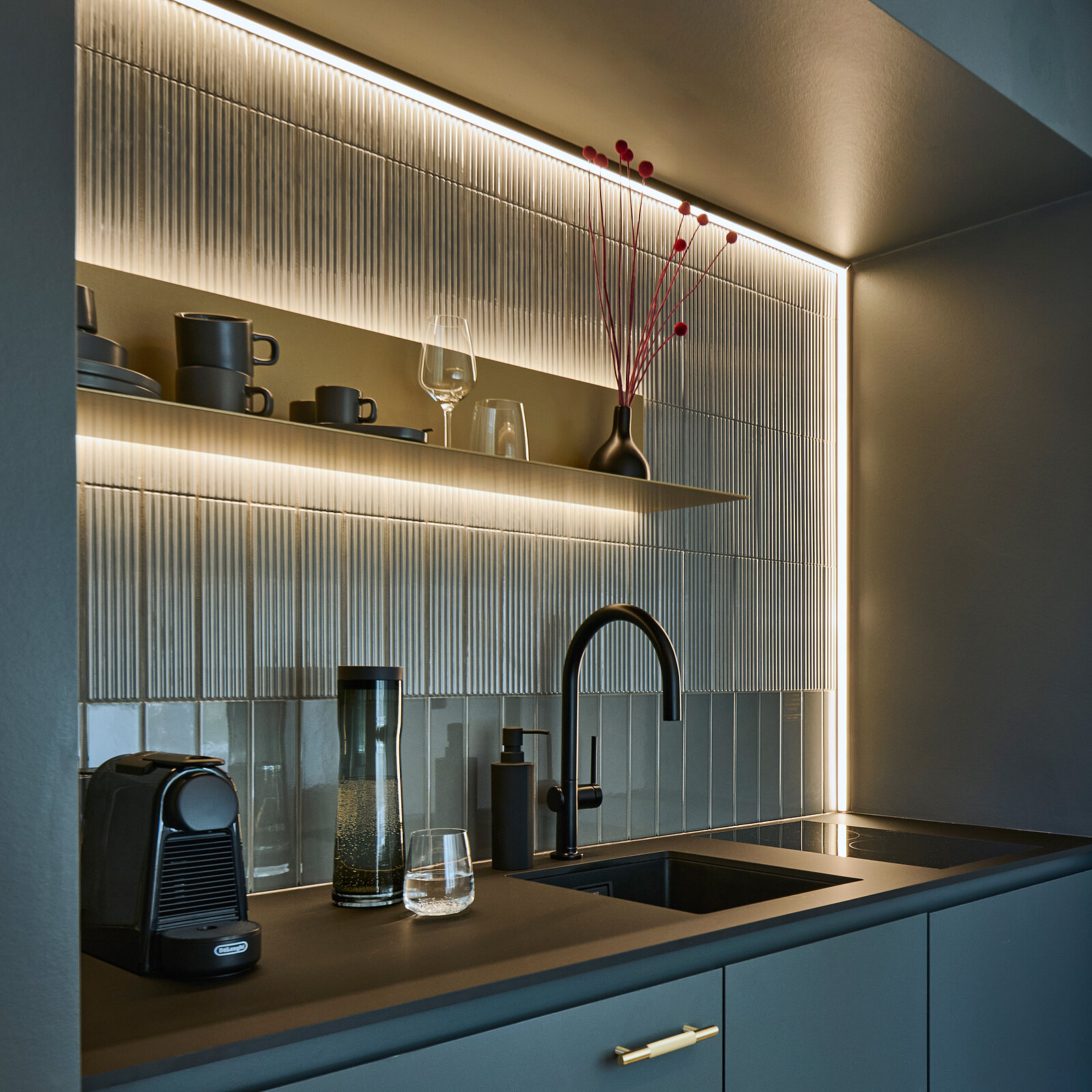 Moderne Küche mit dunkler Arbeitsplatte, integriertem Spülbecken und Wasserhahn. An der Wand sind beleuchtete Regale mit Tassen, Gläsern und dekorativen Elementen. Links steht eine Kaffeemaschine, daneben ein Wasserglas und eine Vase mit roten Zweigen. Dezente Beleuchtung schafft eine stimmungsvolle Atmosphäre.