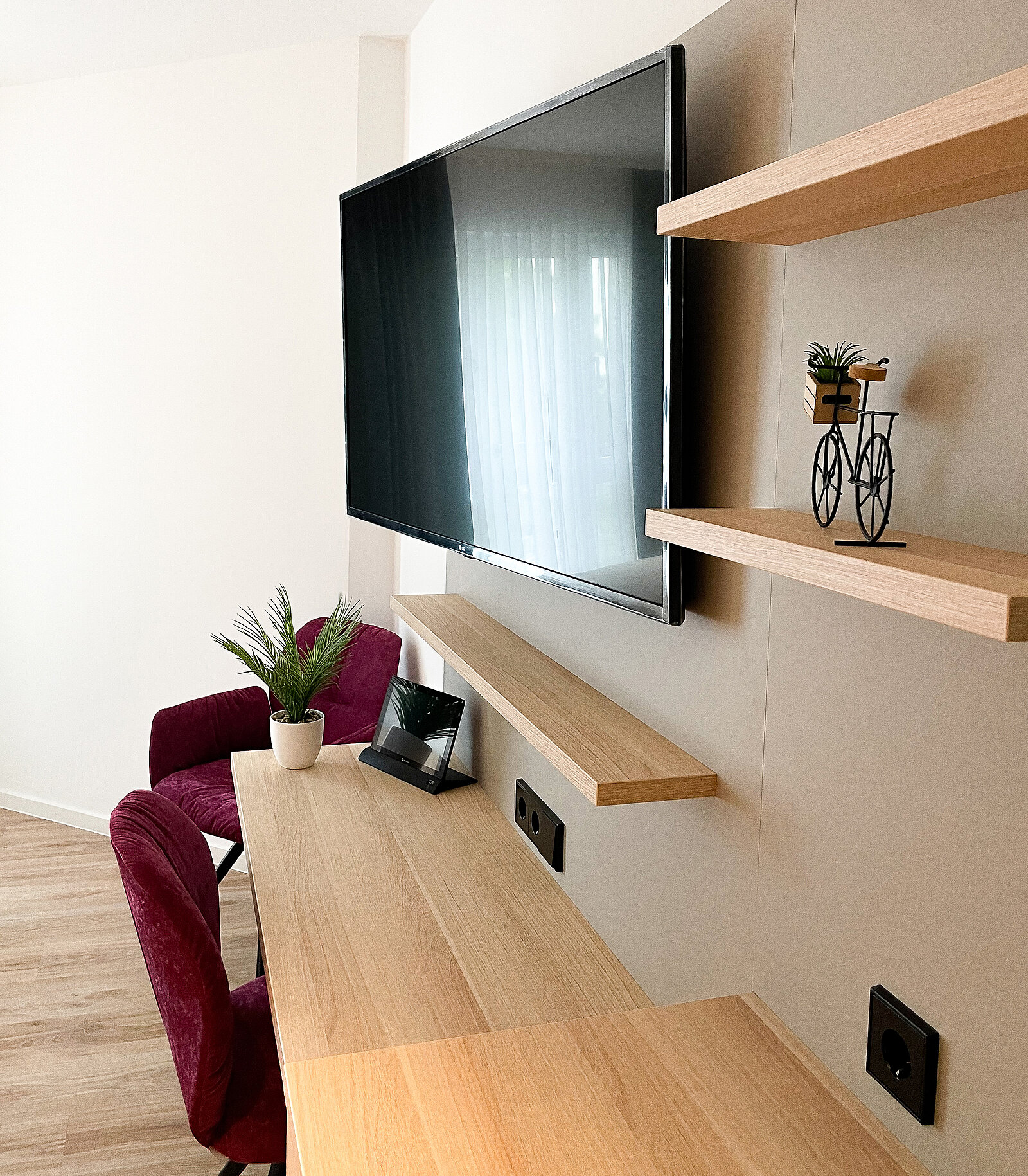 Ein moderner Raum mit einem großen Flachbildfernseher an der Wand, einem Schreibtisch aus Holz mit einem Tablet und einer Pflanze darauf. Es gibt zwei Holzregale mit Dekorationen, darunter eine kleine Fahrradfigur und eine Topfpflanze. Im Hintergrund ein weinroter Stuhl.
