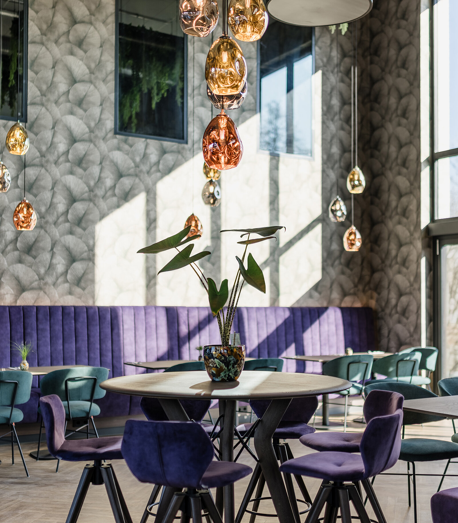 Ein elegantes Café mit hohen Decken, lila Polstermöbeln und hängenden, bunten Lampen. Ein runder Tisch mit einer dekorativen Pflanze steht im Vordergrund. Sonnenlicht fällt durch große Fenster ein.