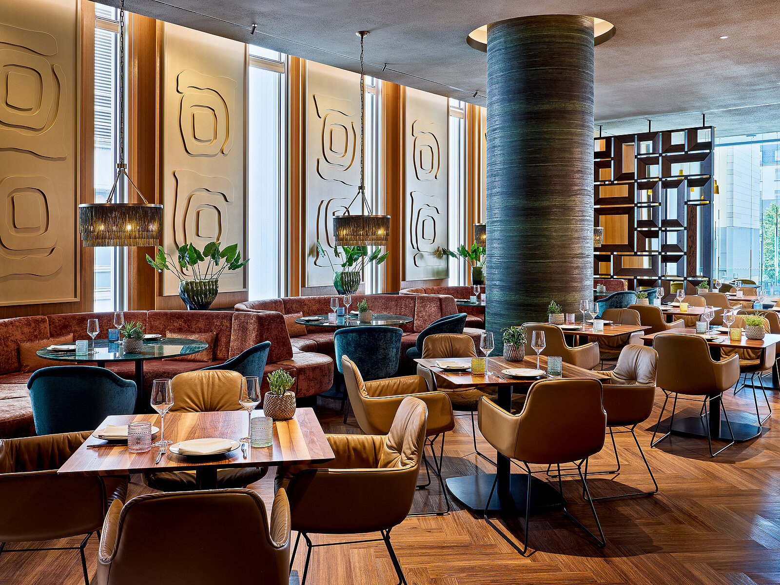 Elegantes Restaurant mit moderner Einrichtung, runden Tischen und gepolsterten Stühlen. Die Wände sind mit abstrakter Kunst dekoriert. Große Fenster sorgen für viel Tageslicht. Pflanzen und stilvolle Beleuchtung ergänzen das Ambiente.
