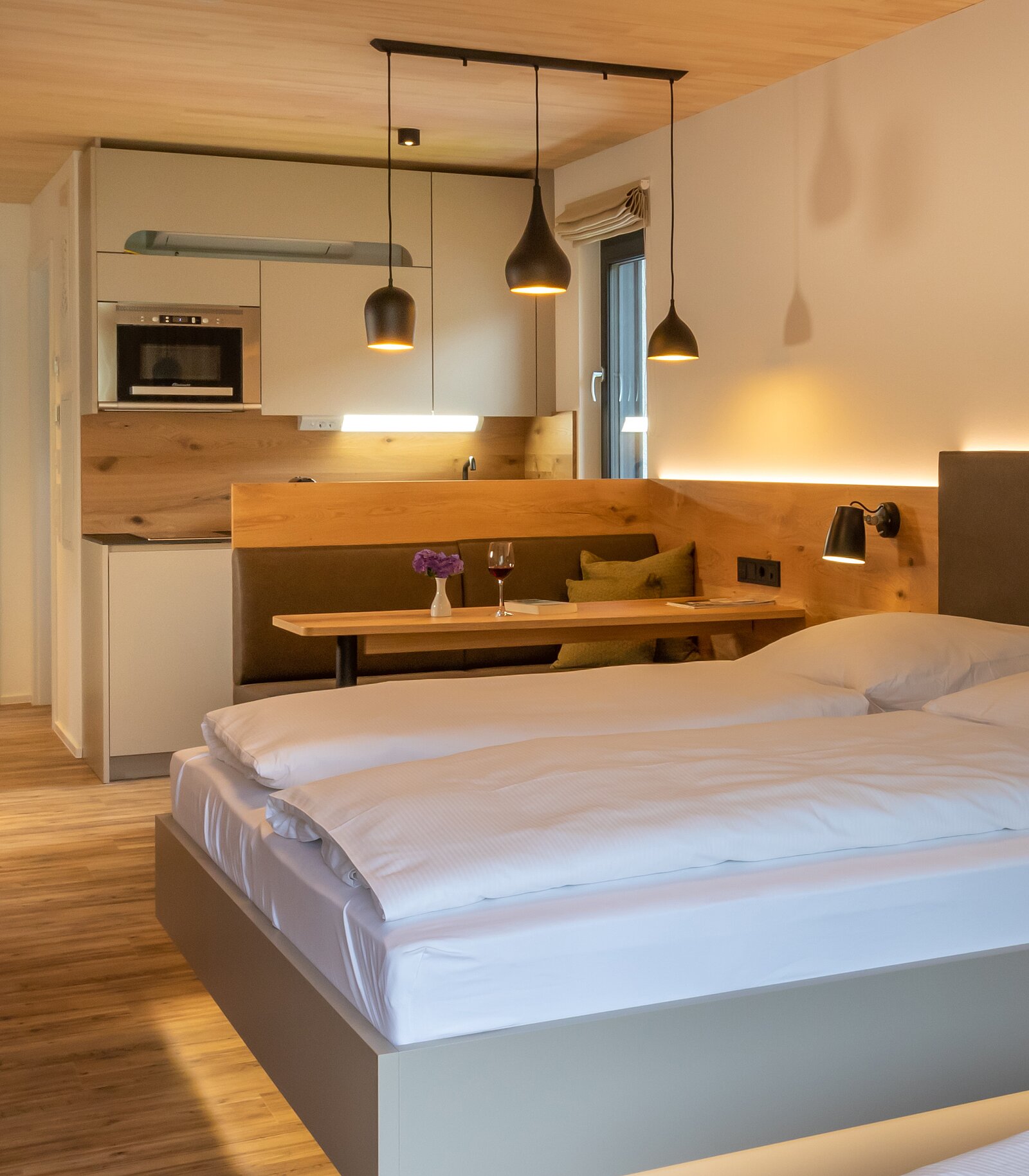 Ein modernes, helles Zimmer mit einem Doppelbett, einer kleinen Küchenecke und einem Esstisch. Die Wände sind in warmen Farben gehalten, und die Beleuchtung wird durch hängende Leuchten ergänzt. Ein Fenster lässt Tageslicht herein, und auf dem Tisch stehen Blumen und Gläser.