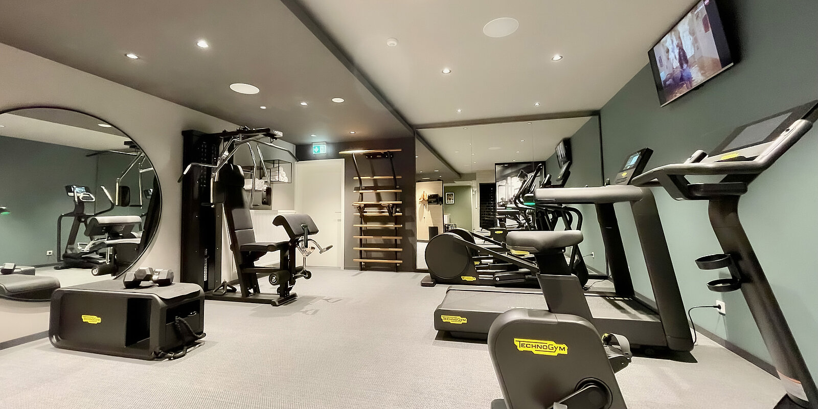 Ein modernes Fitnessstudio mit verschiedenen Geräten. Zu sehen sind ein Laufband, ein Krafttrainingsgerät, ein Ergometer und ein spiegelnder Bereich. Die Wände sind in gedämpften Farben gehalten, und ein Fernseher ist an der Wand montiert.
