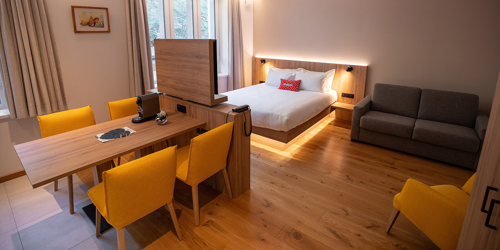 Ein modernes Hotelzimmer mit einem großen Doppelbett, einem grauen Sofa und einem Esstisch mit vier gelben Stühlen. Helle Holzböden und warmes Beleuchtung sorgen für eine einladende Atmosphäre. Im Hintergrund sind große Fenster mit Vorhängen sichtbar.