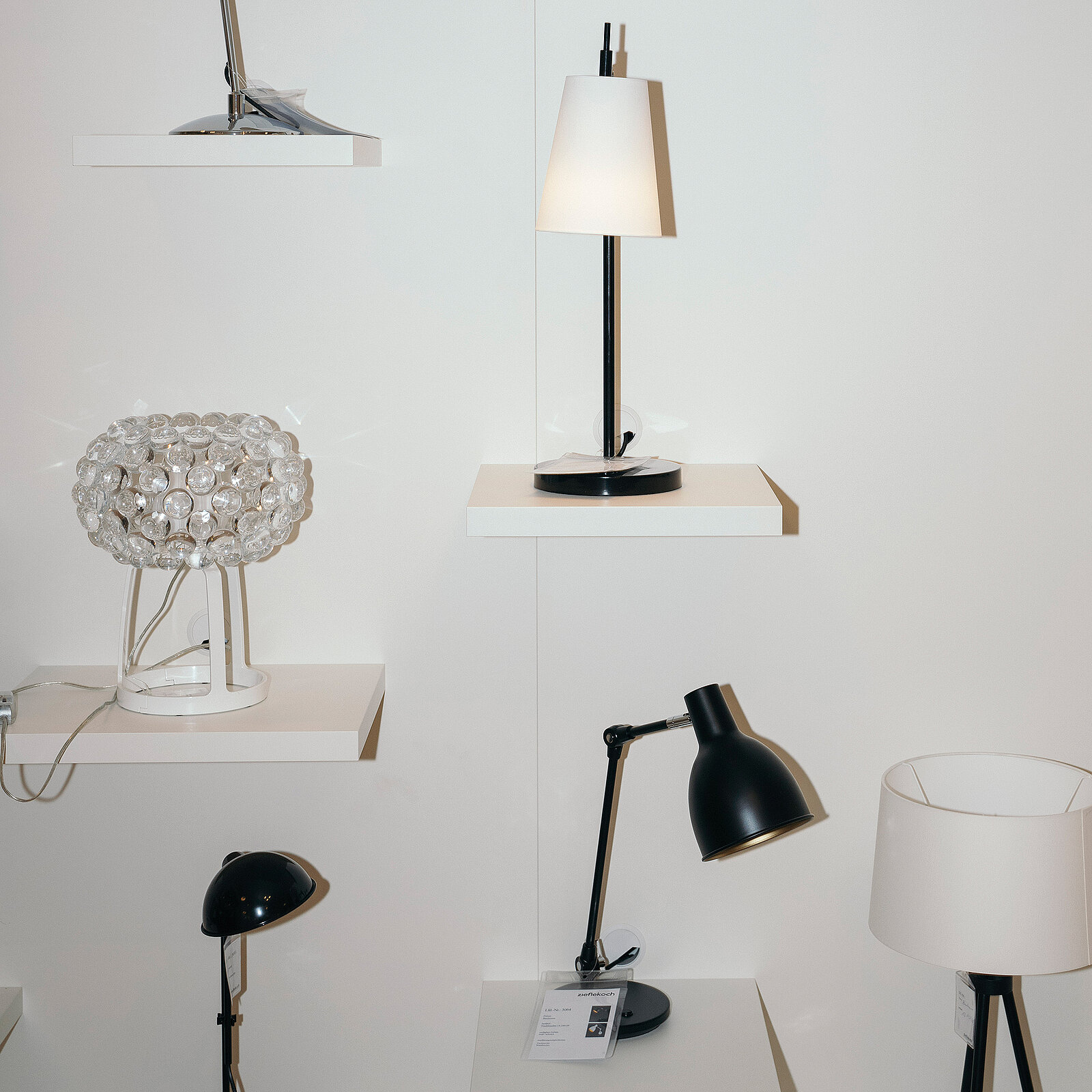 Das Bild zeigt mehrere Lampen, die auf weißen Regalen angeordnet sind. Darunter befinden sich eine klare Glaslampe, eine schwarze Tischlampe und eine Stehlampe mit einem weißen Lampenschirm auf einem schwarzen Fuß. Die Anordnung fokussiert sich auf Beleuchtungskörper in verschiedenen Stilen.