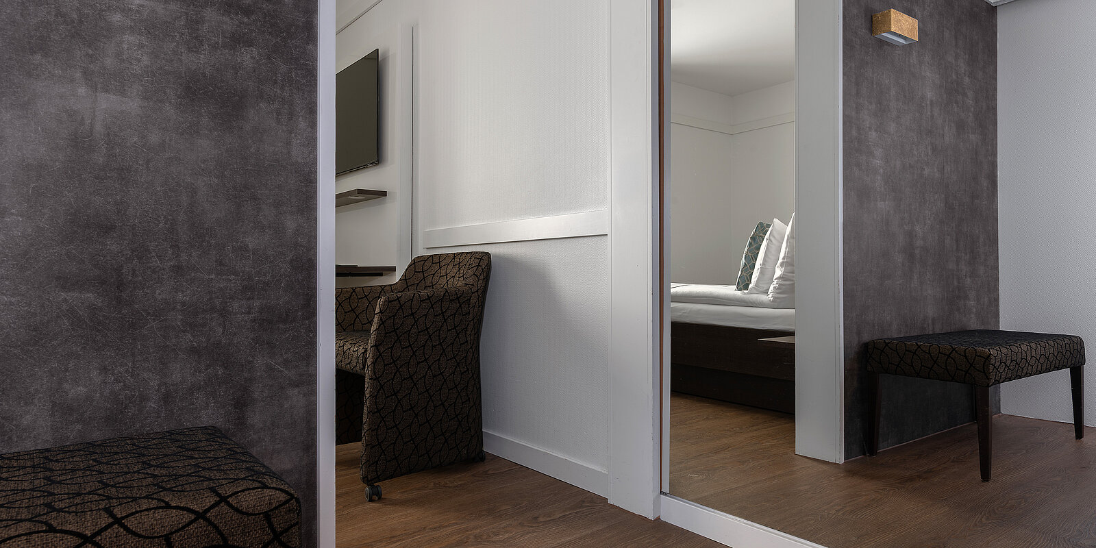 Ein modernes Hotelzimmer mit einem gemütlichen Doppelbett, grauen Wänden und Holzboden. Ein Sessel und eine Bank mit dunklem Stoffmuster sind sichtbar. Ein Spiegel an der Wand erweitert den Raum optisch.
