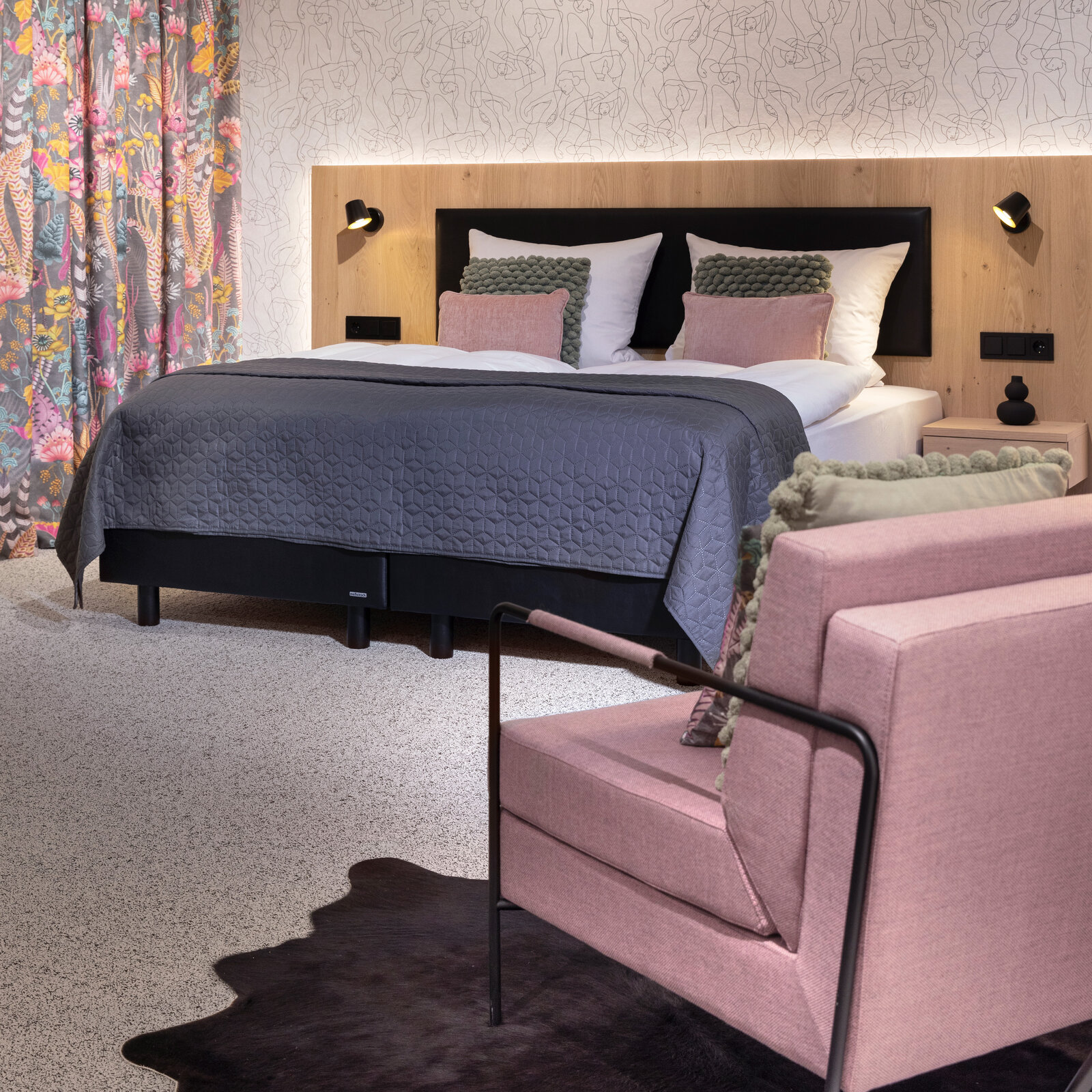 zieflekoch musterzimmer zenit, modernes Schlafzimmer mit einem Boxspring-Doppelbett mit Rückwand in Holzoptik, einem rosafarbenen Sessel und einer abstrakten Motivtapete, der Teppichboden und ein dunkler Teppich verleihen dem Raum Wärme.