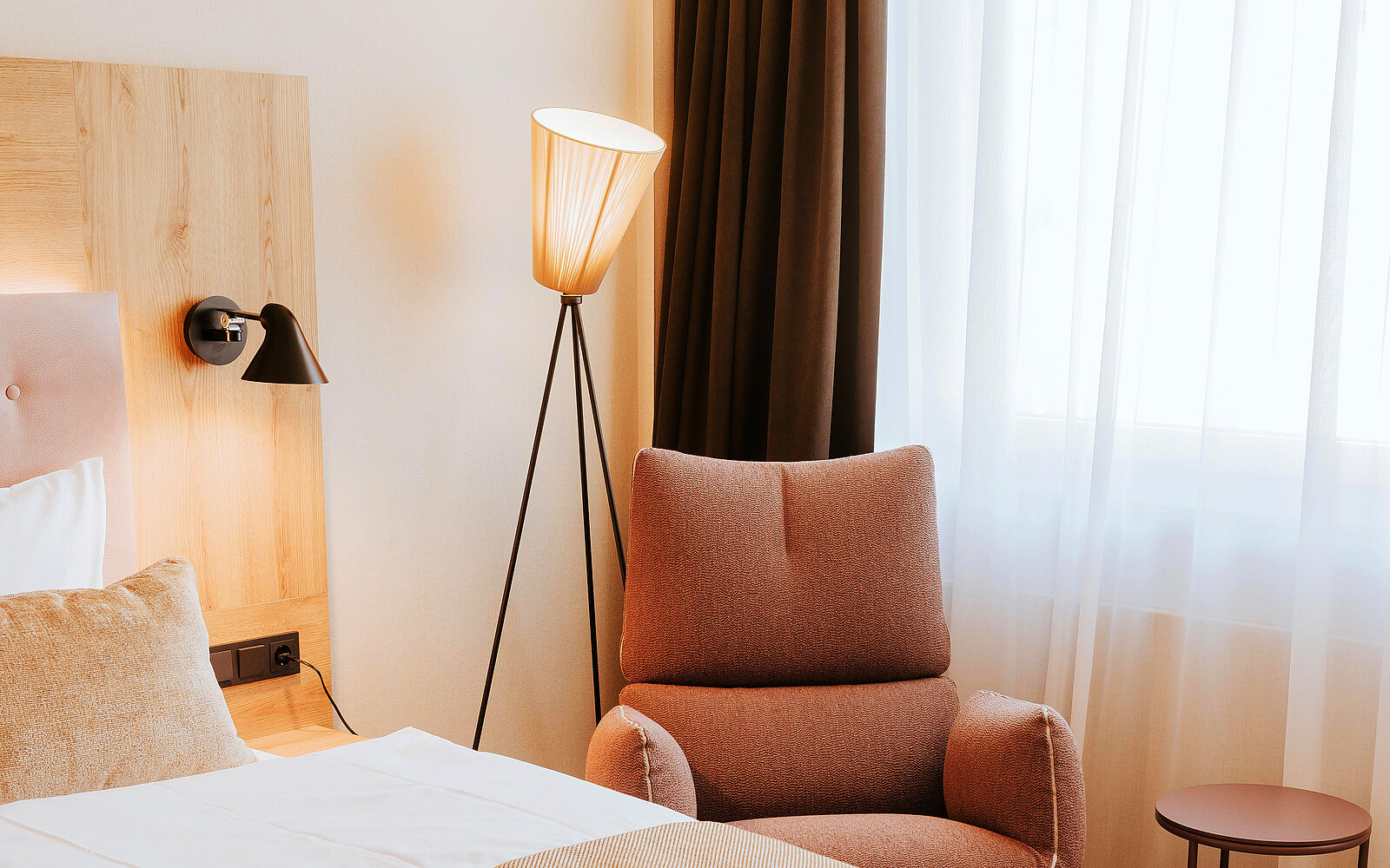Ein gemütliches Hotelzimmer mit einem Bett, einer modernen Stehlampe und einem beigen Sessel. Große Fenster lassen viel Tageslicht herein, während ein kleiner Tisch in der Nähe steht. Holz- und Textilmaterialien schaffen eine warme Atmosphäre.