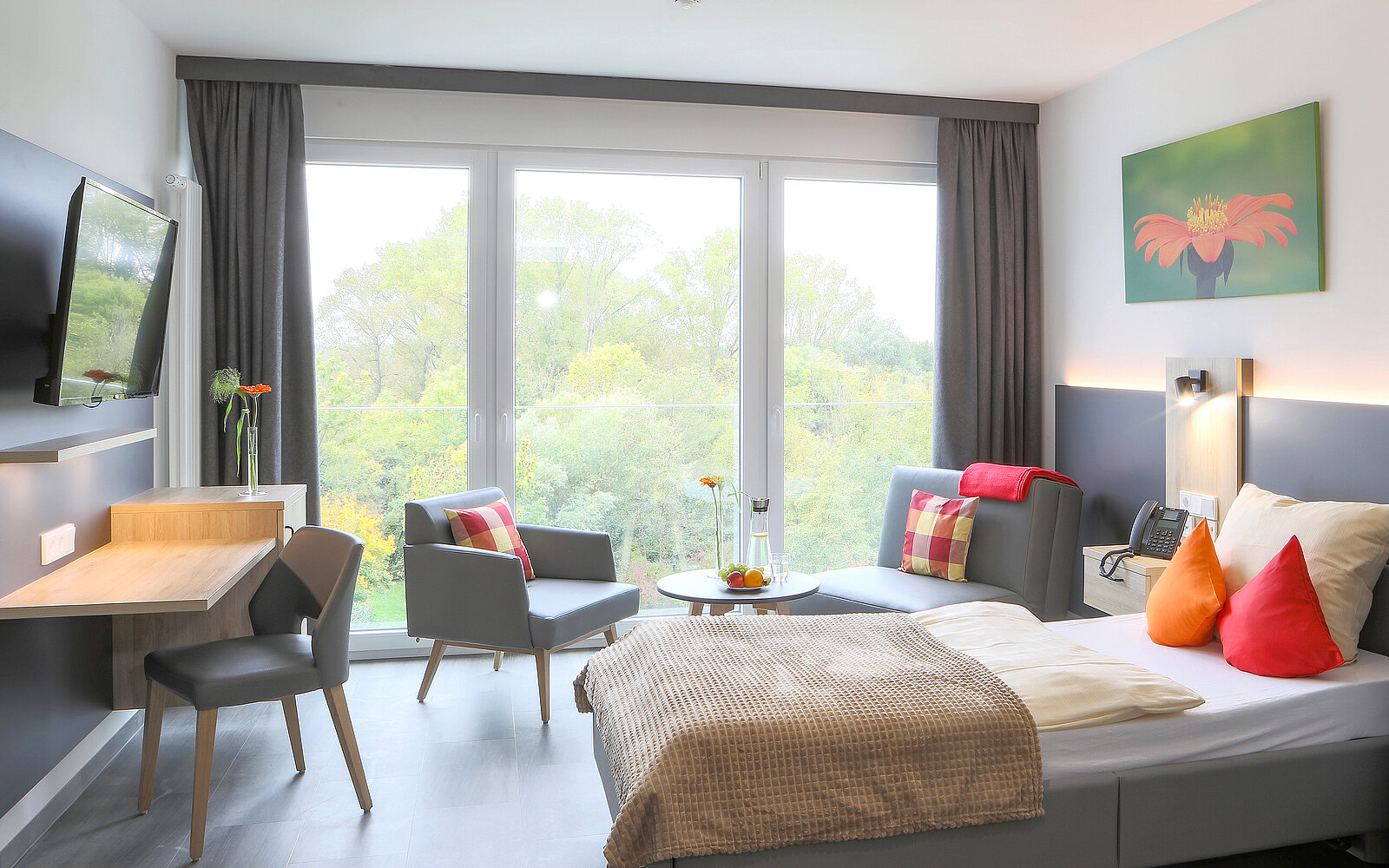 Ein helles Hotelzimmer mit einem Einzelbett, einem Schreibtisch und zwei Stühlen. Vor dem Fenster sind Pflanzen und eine Aussicht auf Bäume sichtbar. An der Wand hängt ein Bild mit einem Flamingo. Der Raum ist modern und einladend gestaltet.