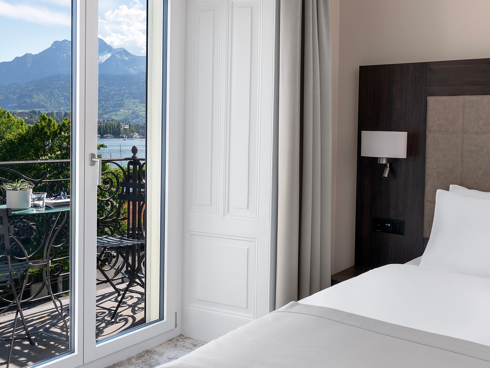 Ein Hotelzimmer mit einem großen Fenster, das auf einen Balkon führt. Vom Balkon aus sieht man eine malerische Aussicht auf einen See und Berge im Hintergrund. Ein Bett steht rechts im Raum, und auf dem Balkon gibt es Sitzgelegenheiten.