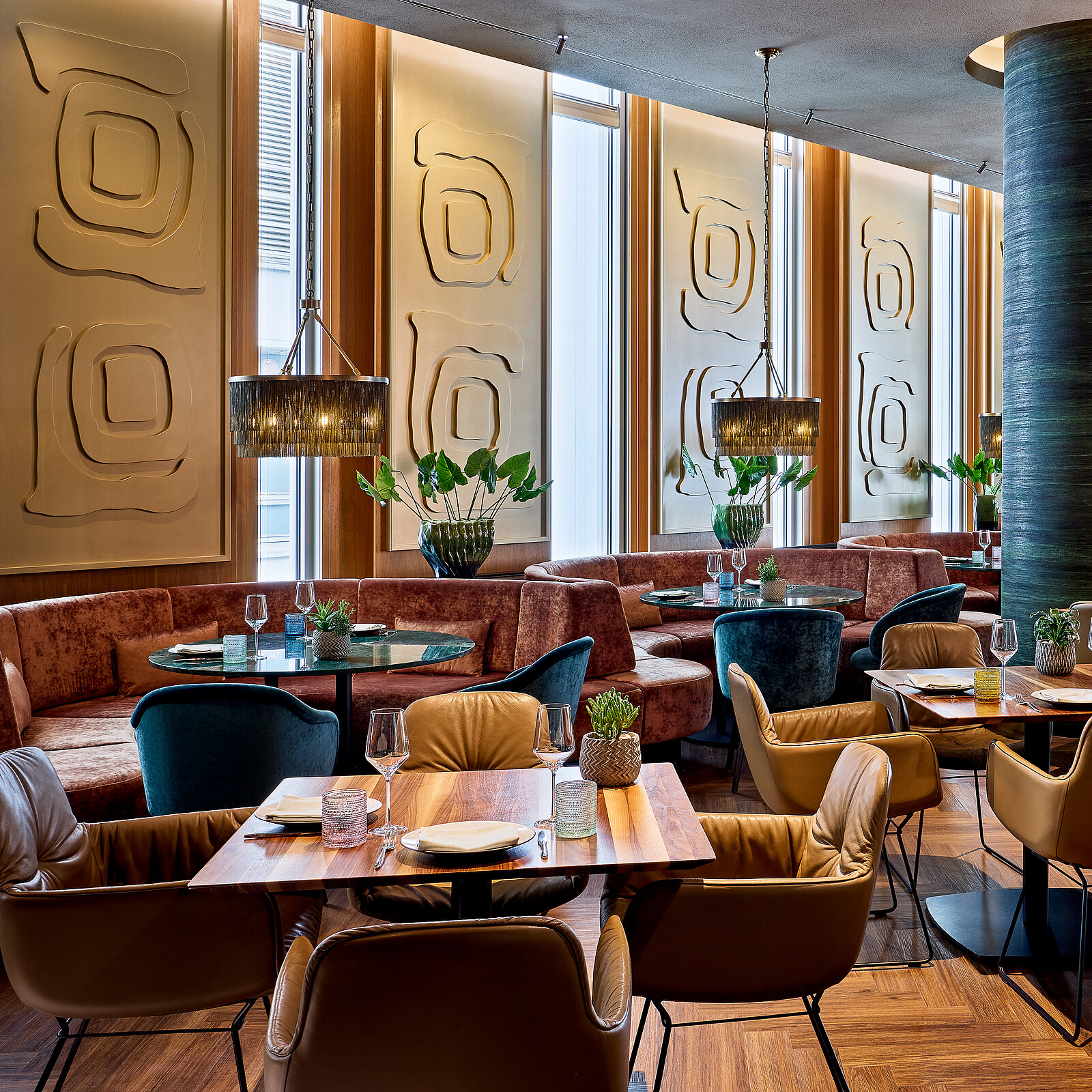 Gemütliches Restaurant mit eleganter, moderner Einrichtung. Runde Tische mit blauen und braunen Sesseln. Dekorative Wandleuchten und Pflanzen sorgen für eine angenehme Atmosphäre. Holzfußboden und stilvolle Wandverzierungen ergänzen das Ambiente.